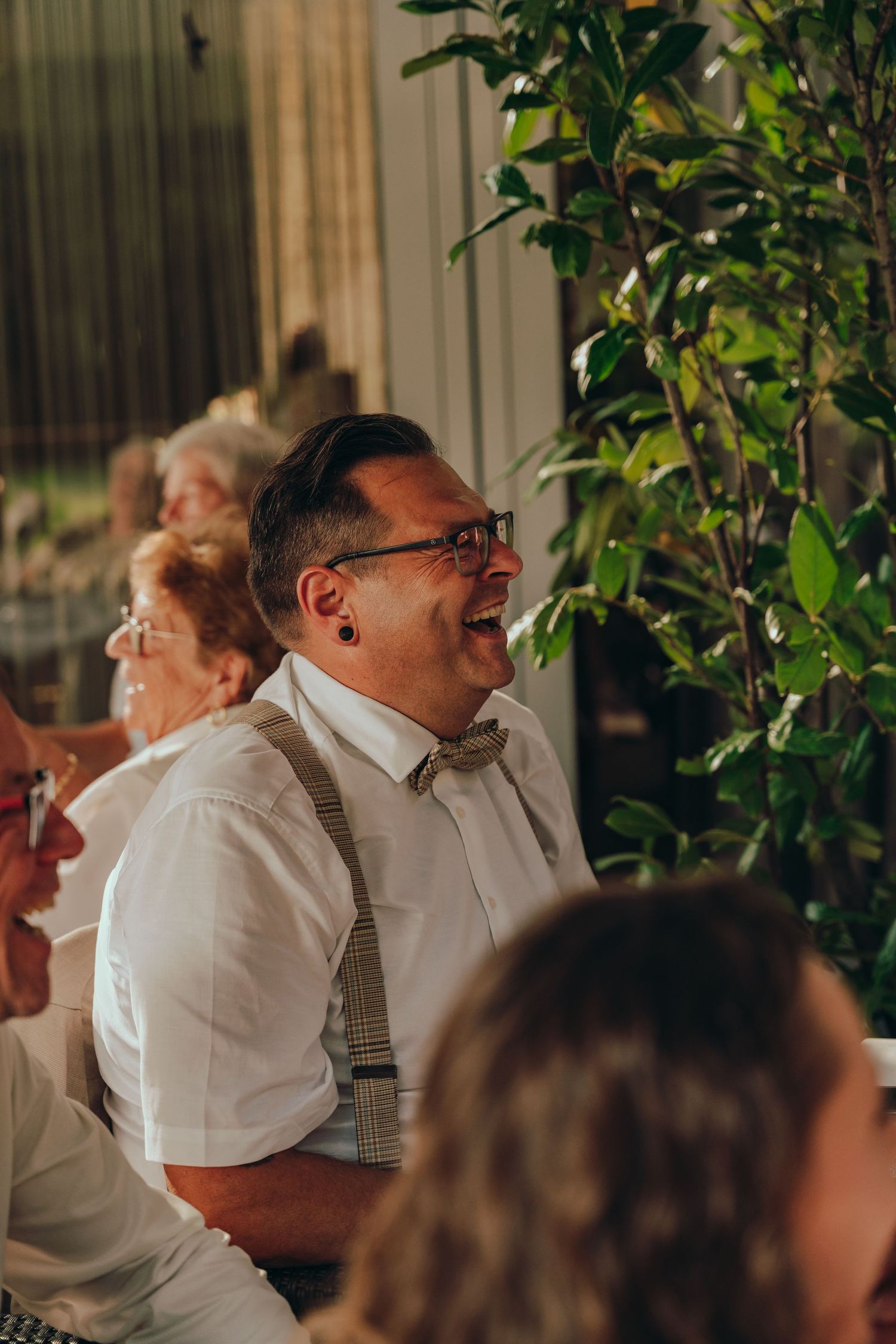 Laughing wedding guest wearing glasses, bow tie and suspenders enjoys reception celebration.