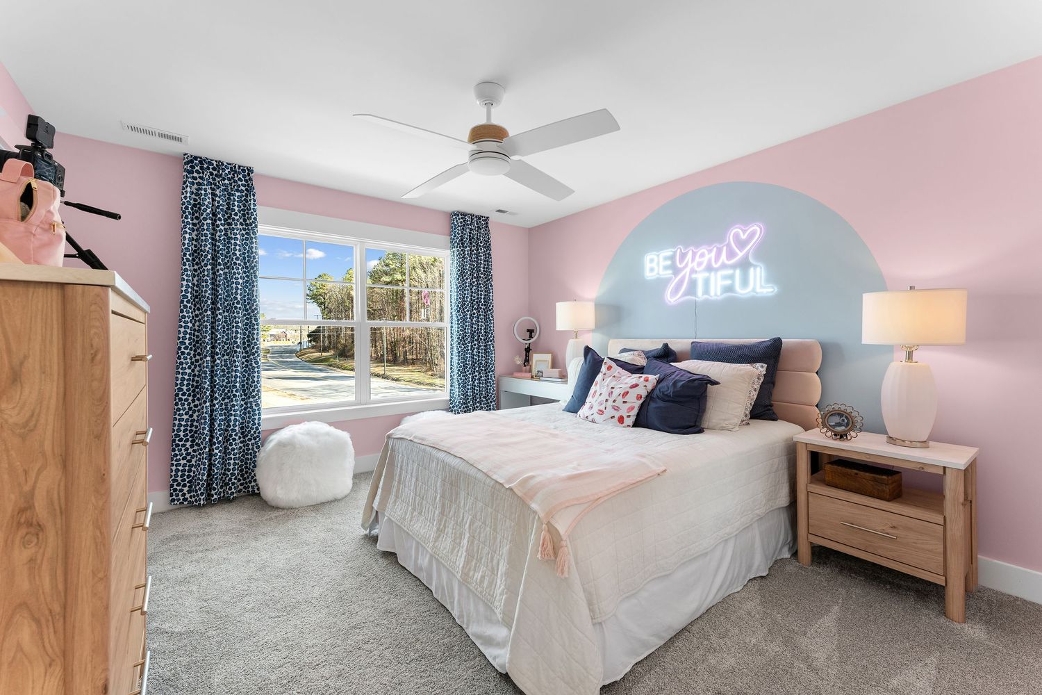 A bright and airy bedroom features pink walls, blue curtains, white bedding, and modern furnishings.
