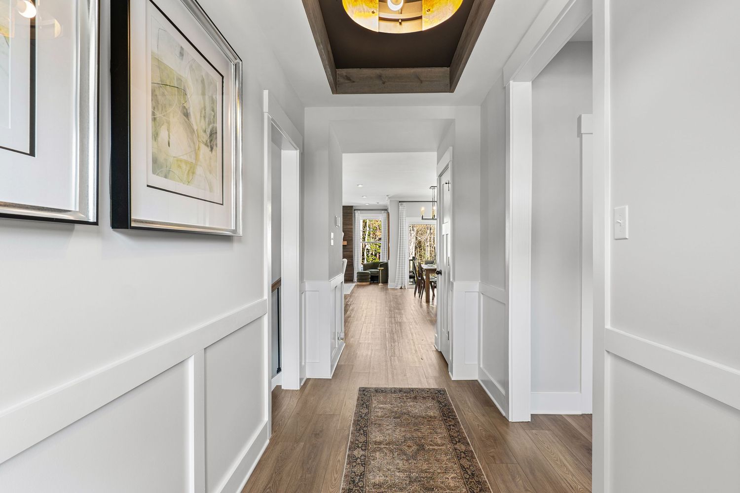 Modern hallway with white wainscoting, hardwood floors, and a brown runner rug leading to a bright interior.