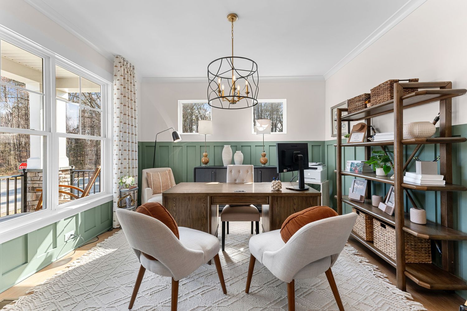 Modern home office with sage green wainscoting, wooden shelving units, and a circular gold chandelier above a wooden desk.