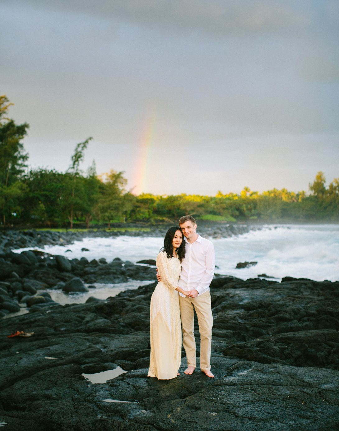 Couple standing on black lava rocks at sunset with a rainbow in the distance at Hilo, Big Island of Hawaii.