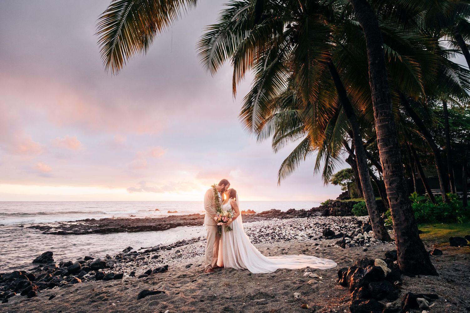 Palm trees frame a romantic beach scene at sunset with rocky coastline in Kona, Big Island Hawaii.