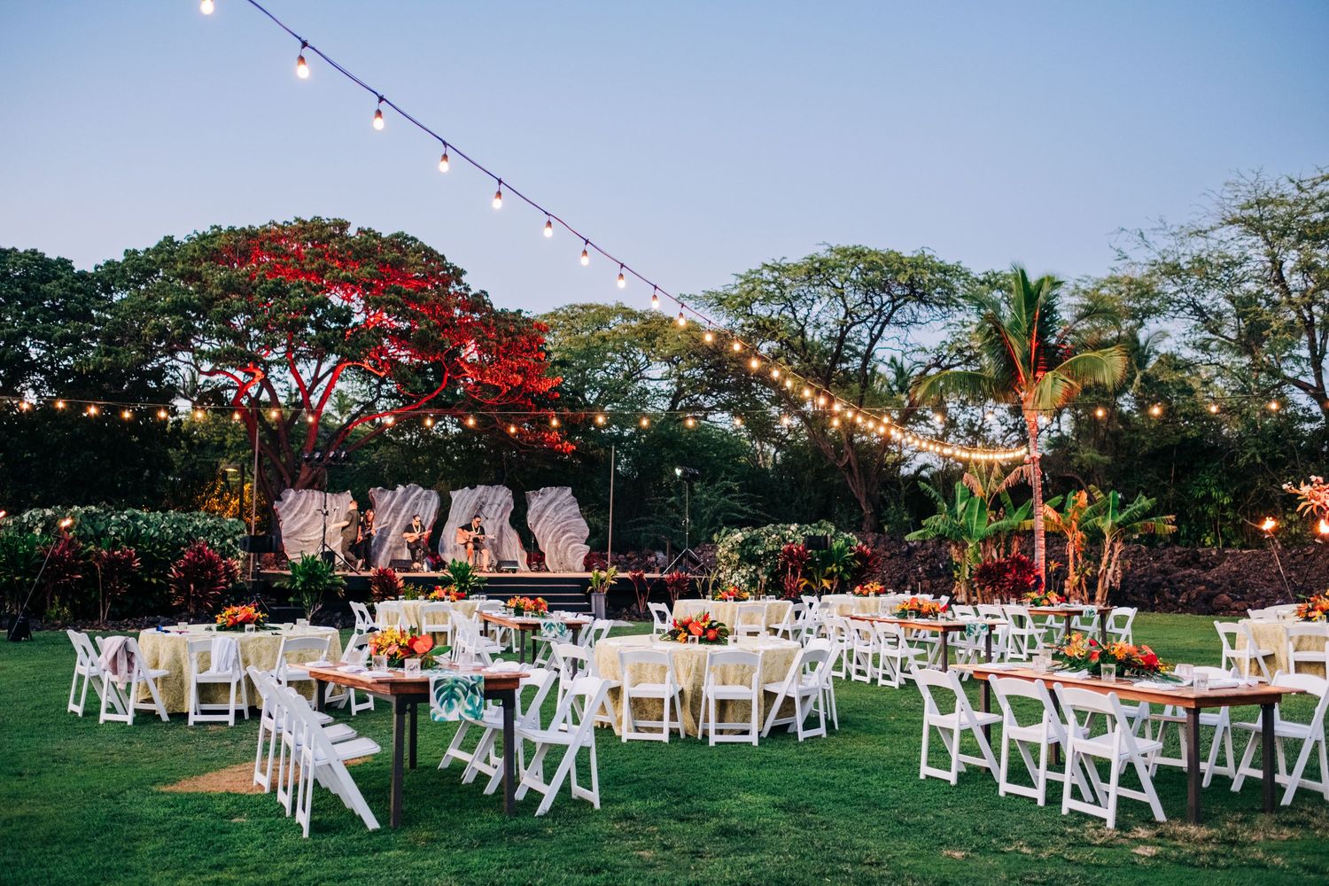 Outdoor wedding reception setup with string lights and white chairs under tropical trees at Mauna Lani Beach Resort.