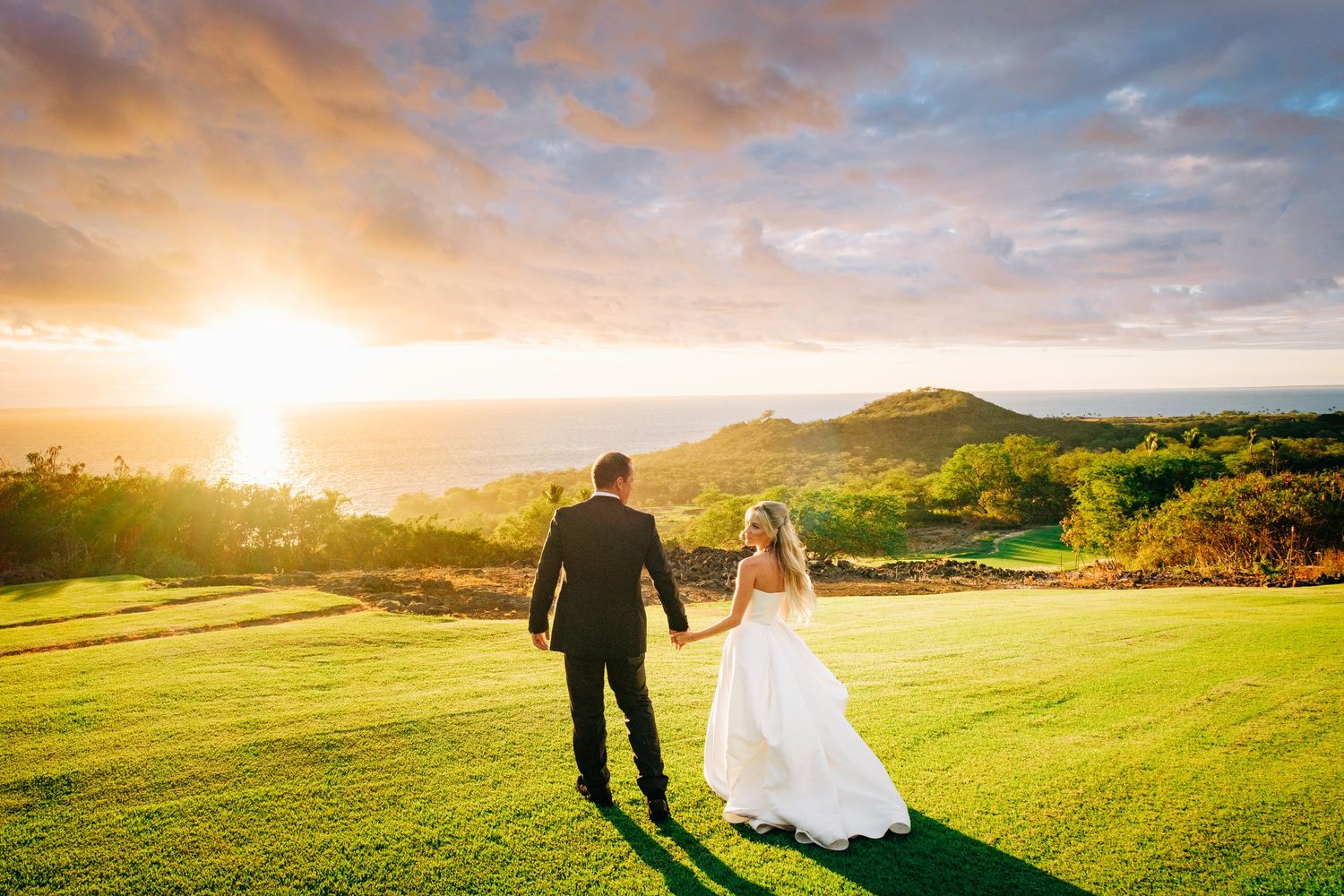 Kohanaiki wedding couple walks across a lush green hilltop meadow at sunset with dramatic mountain views in Kona, Hawaii.