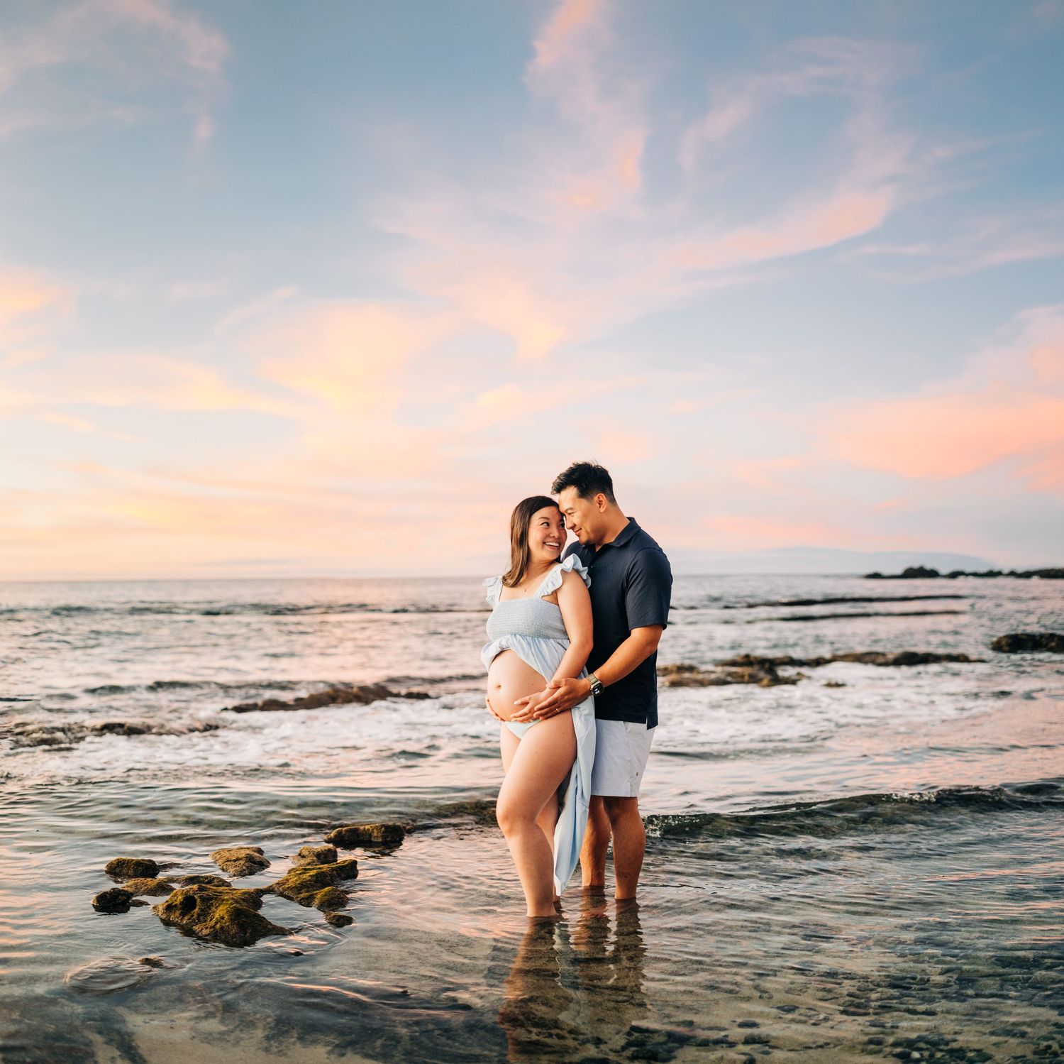 Maternity beach photos with couple standing in shallow ocean water at sunset at Kukio Beach, Kona, Big Island Hawaii.