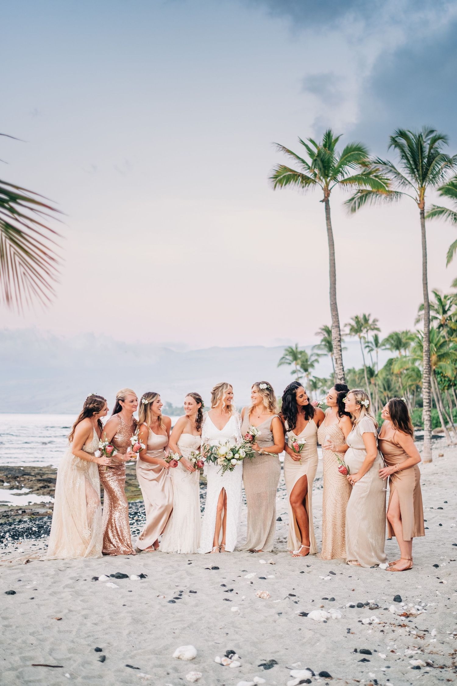 Wedding party in neutral dresses standing on a black sand beach with palm trees at Mauna Lani Beach Resort.
