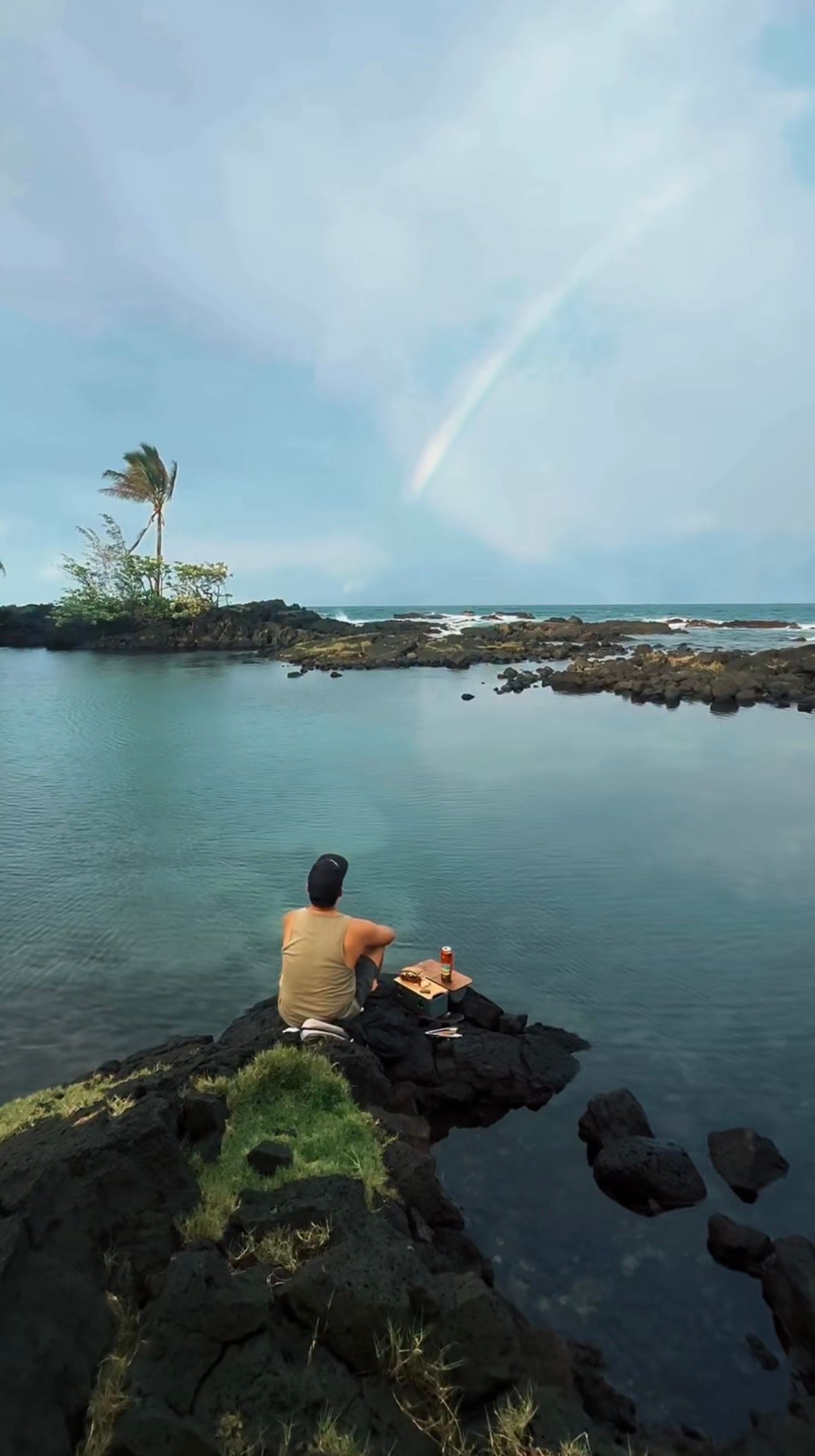 James Rubio sitting on volcanic rocks overlooking tranquil ocean cove in Hawaii.