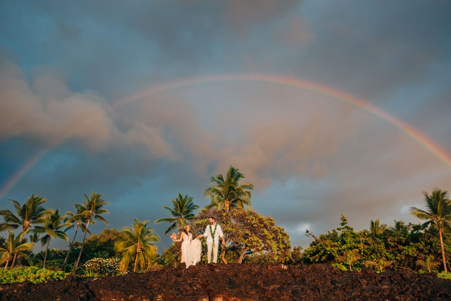 A wedding couple stands silhouetted against a dramatic rainbow and palm trees in Hawaii at Outrigger Kona Resort, Big Island Hawaii.