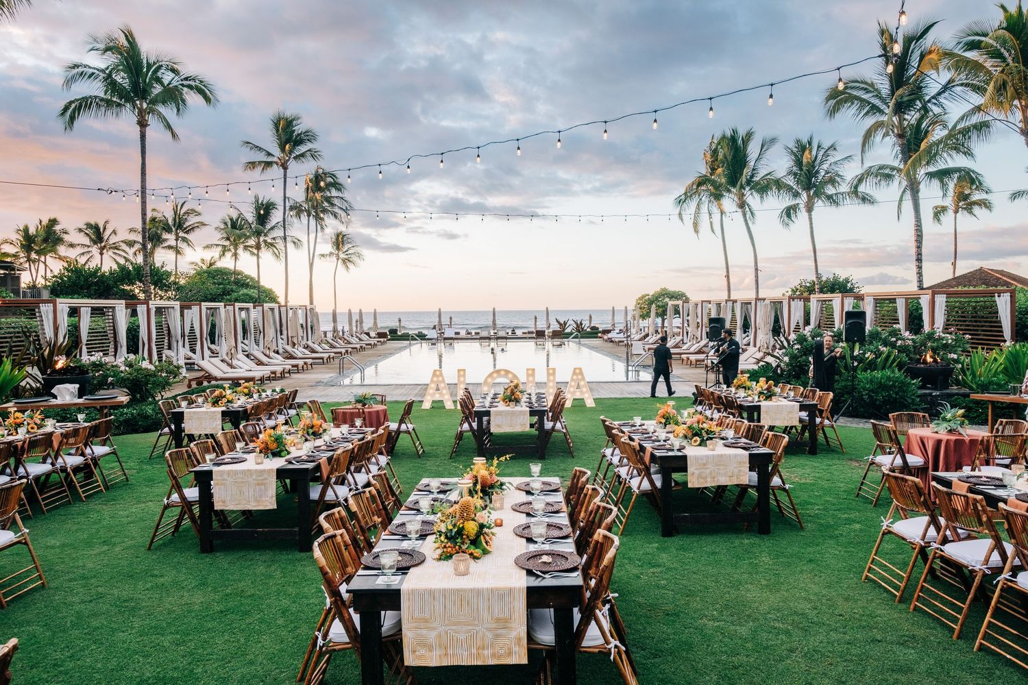 Elegant outdoor wedding reception setup with tables on grass overlooking the ocean at sunset at a Four Seasons Hualalai Event.