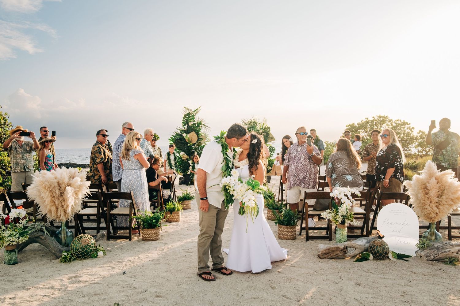 Wedding guests gather on beach for sunset ceremony with wooden chairs at Kona Salt Farm, Big Island of Hawaii.