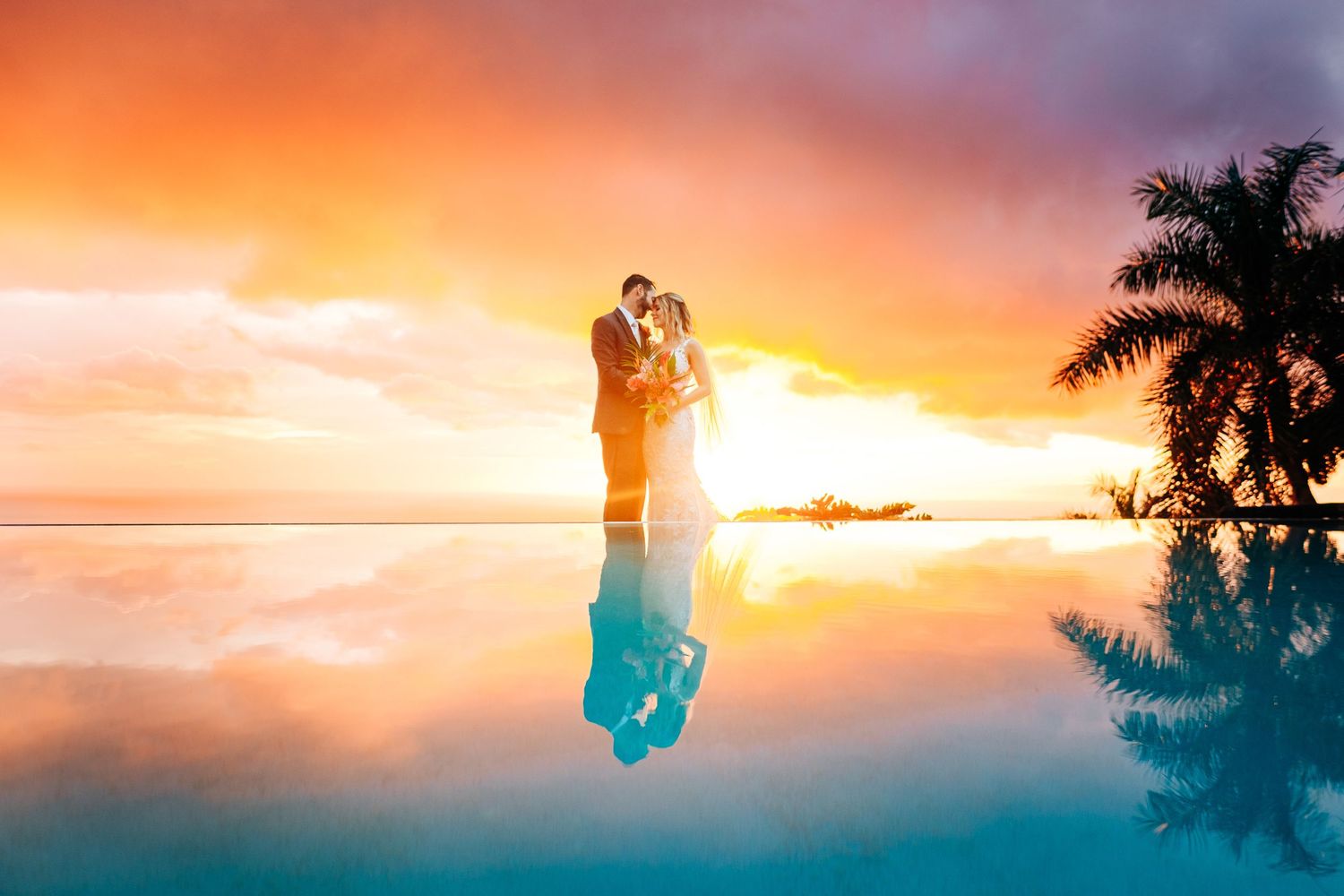 Silhouettes embrace against a vibrant orange and pink Hawaiian sunset with palm trees framing the romantic scene in Kona, Big Island.