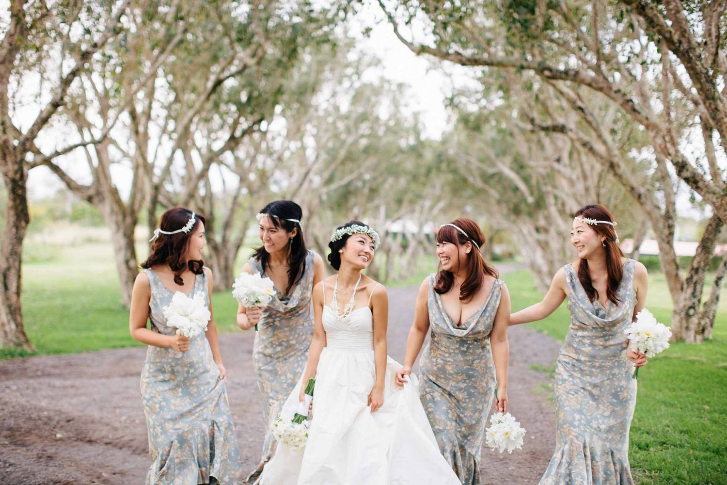 Bridesmaids in light colored dresses walking together through a sunlit wooded area at Puakea Ranch, Big Island of Hawaii.