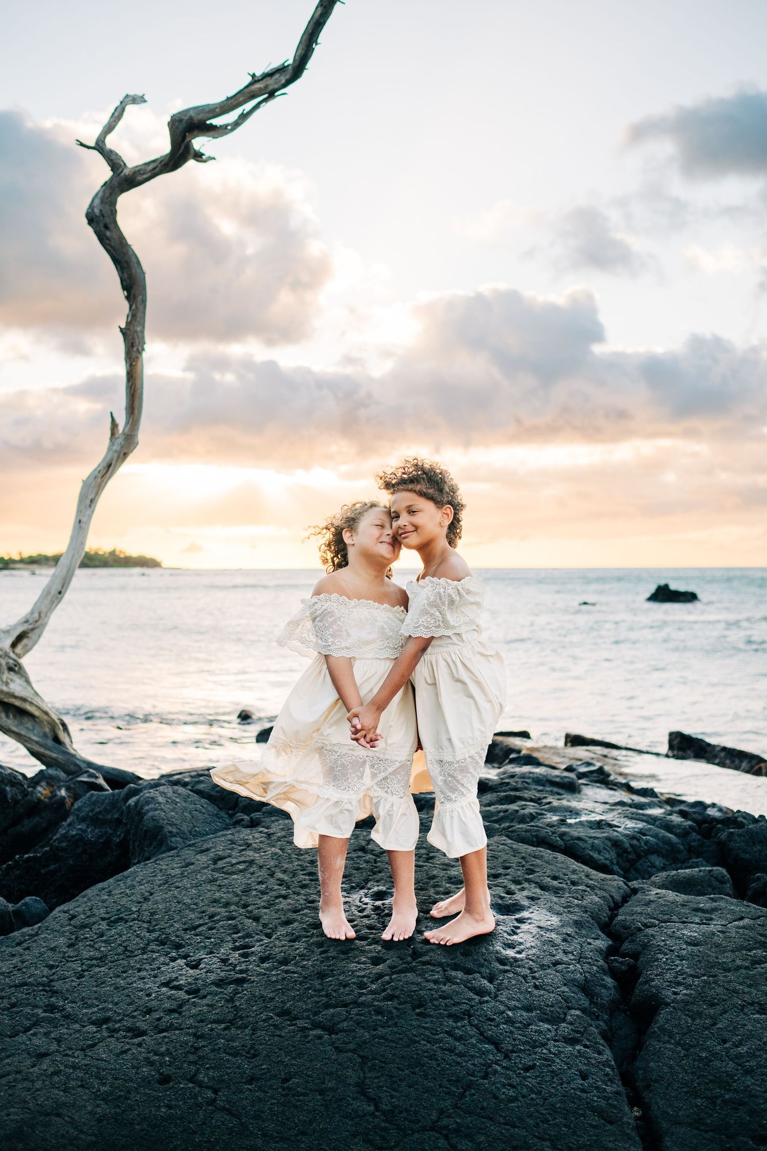 Children embrace while standing on volcanic rocks at sunset near the ocean with a twisted tree branch at A-bay Waikoloa, Big Island Hawaii.