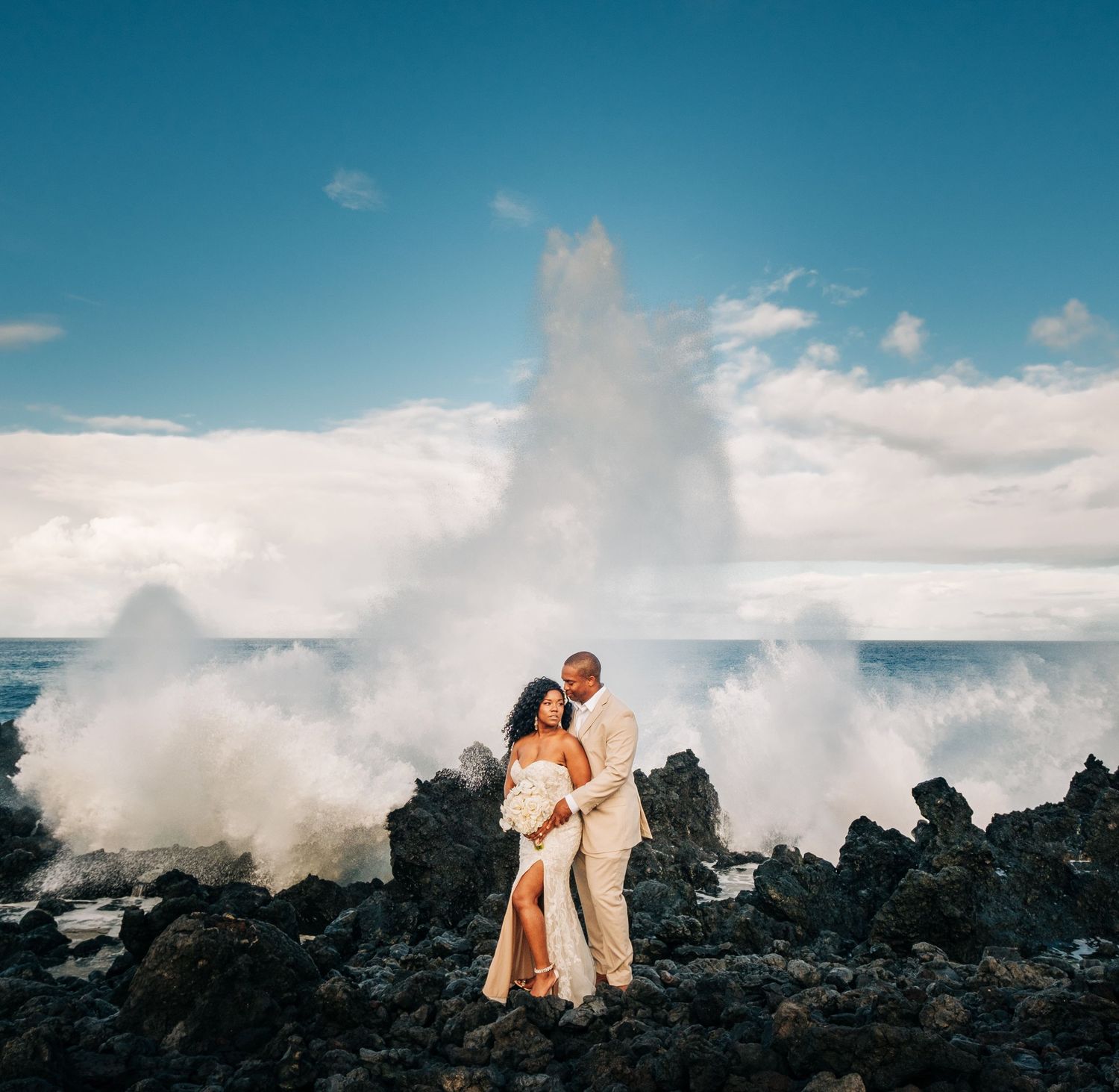 Dramatic ocean waves crash against volcanic rocks as couple poses for wedding photos in Kona, Big Island Hawaii.