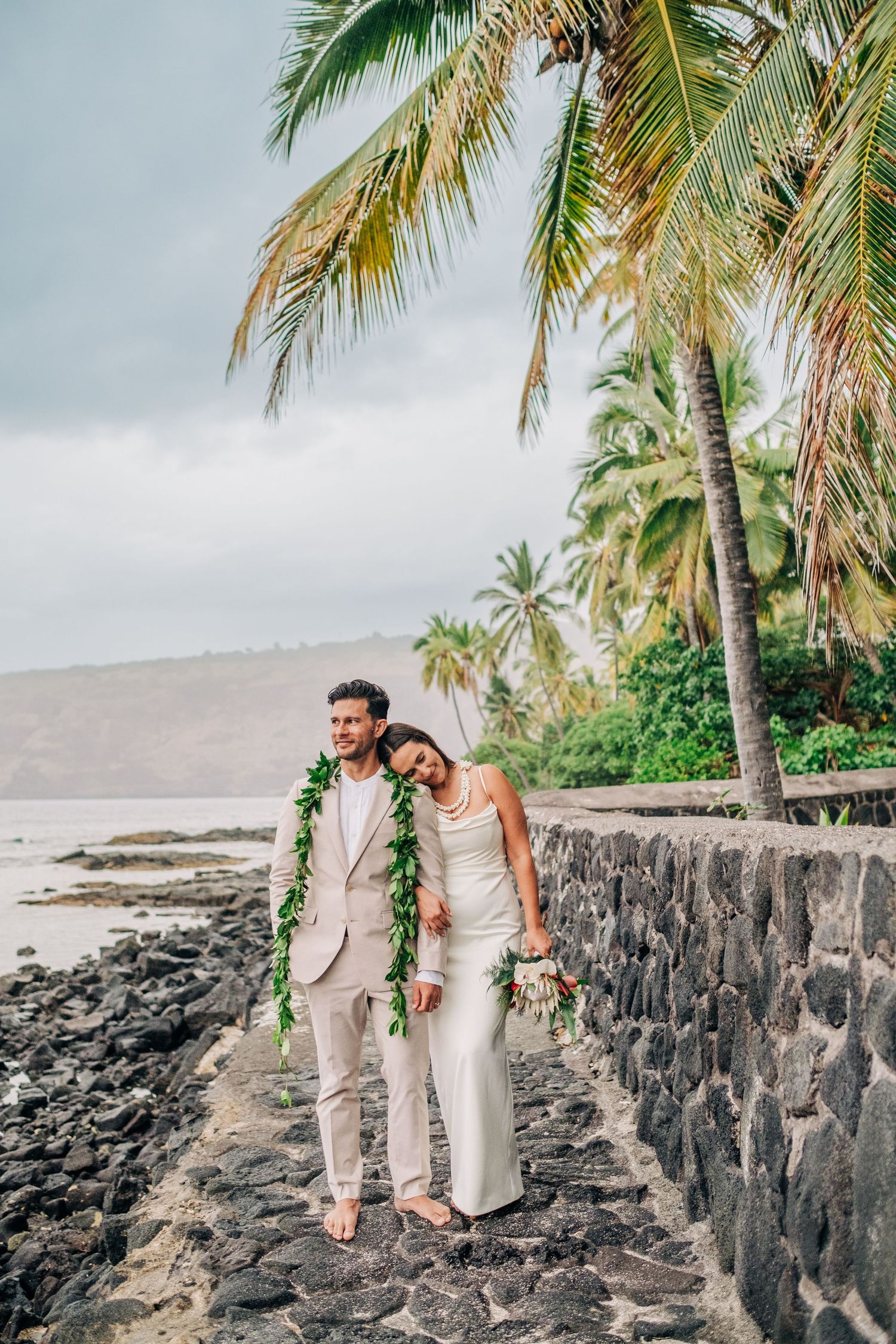 A couple walks along a rocky beach coastline with palm trees and ocean views in Kona, Big Island of Hawaii.