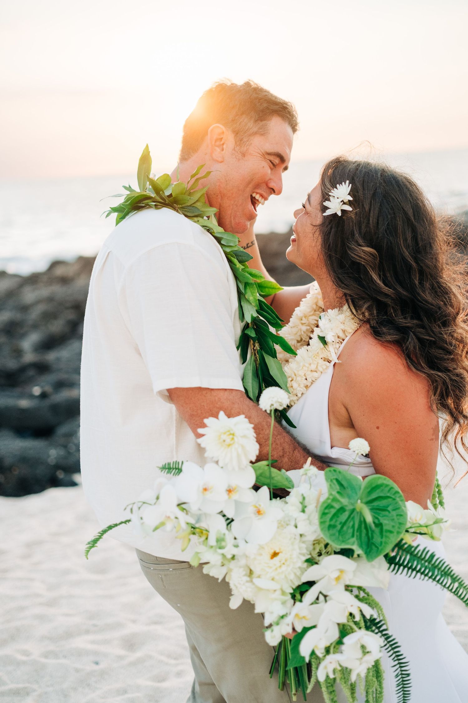 Couple in white wedding attire embrace while wearing traditional Hawaiian leis on a beach at Kona Salt Farm, Big Island of Hawaii.