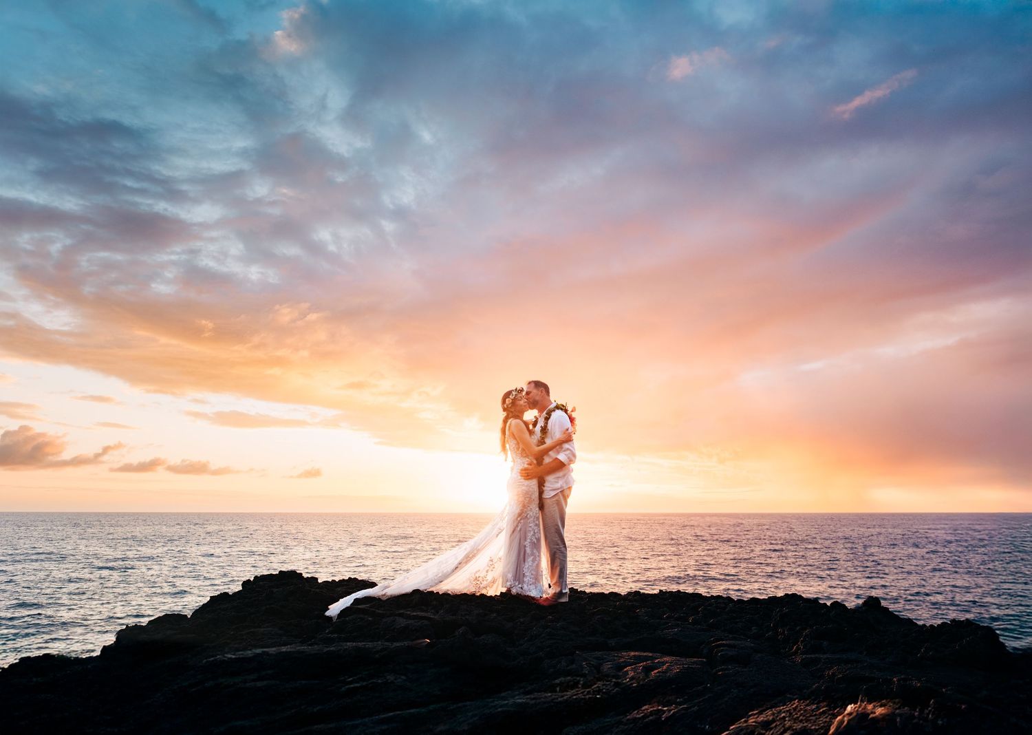 Newlyweds embrace on rocky coastline during golden sunset over the Pacific Ocean at Outrigger Kona Resort, Big Island Hawaii.