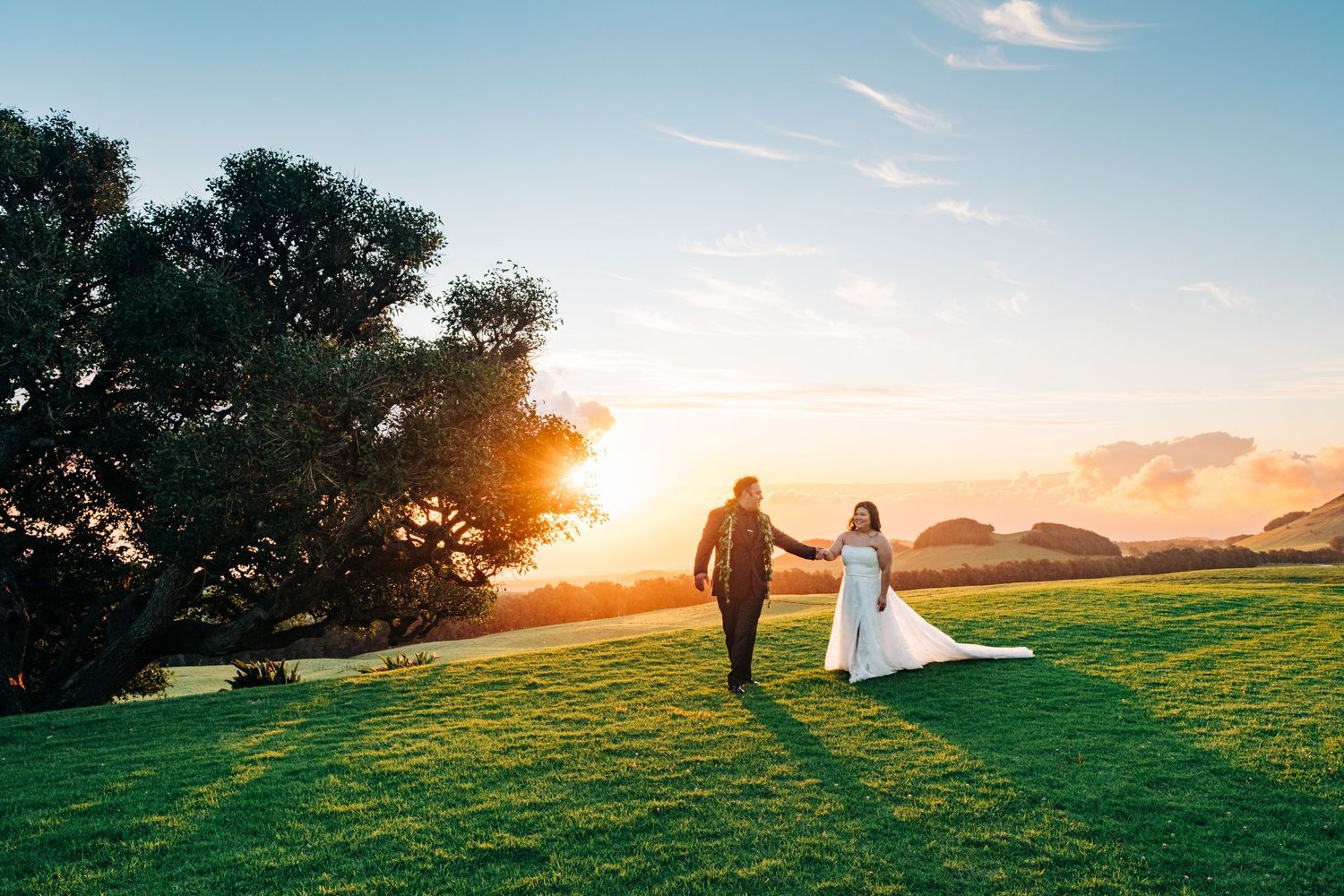 Wedding couple walks hand in hand across grassy hill during golden sunset at Kahua Ranch, Big Island Hawaii.