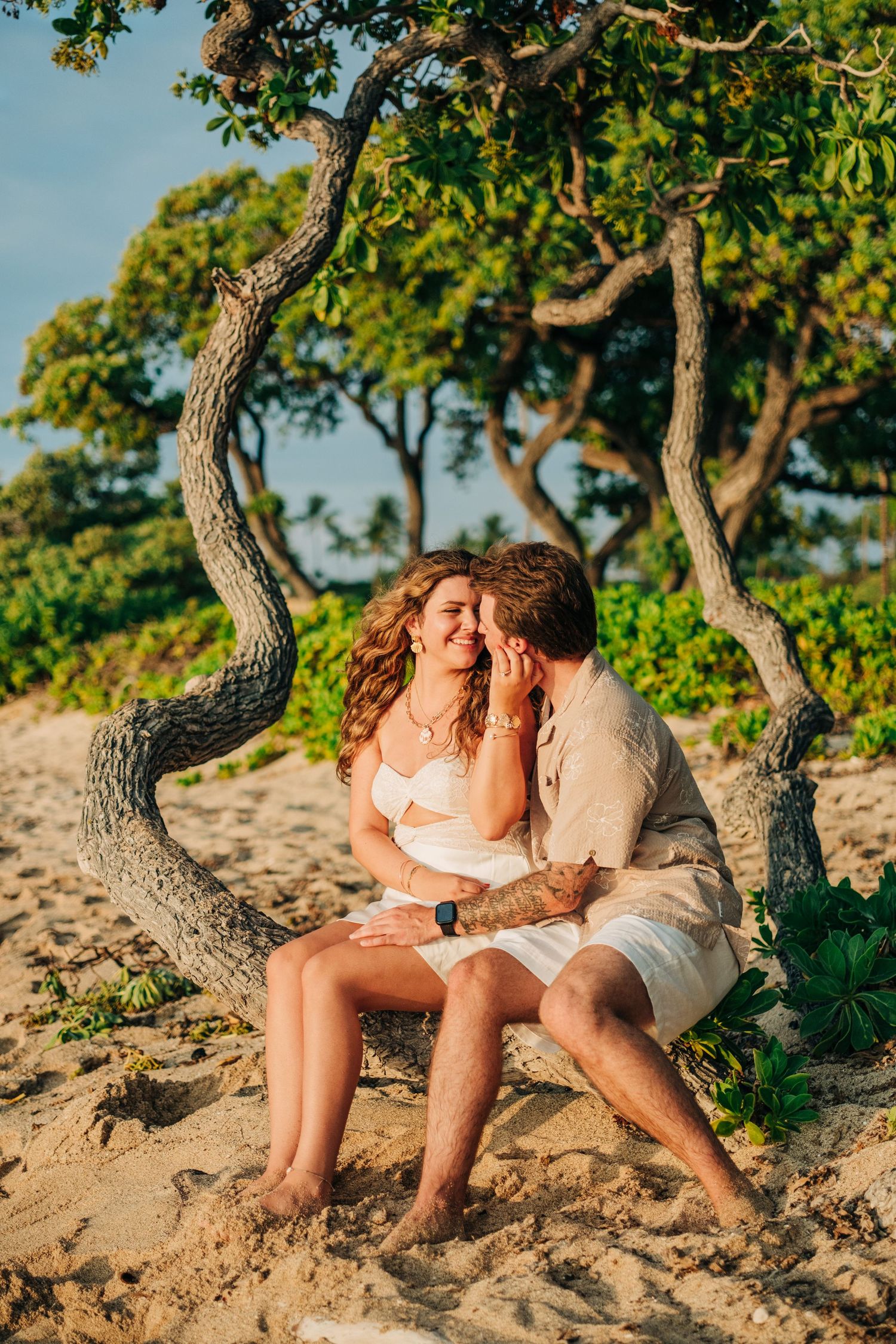 Couple shares intimate moment under twisted beach trees during golden hour photography session at Kukio Beach, Kona, Big Island of Hawaii.