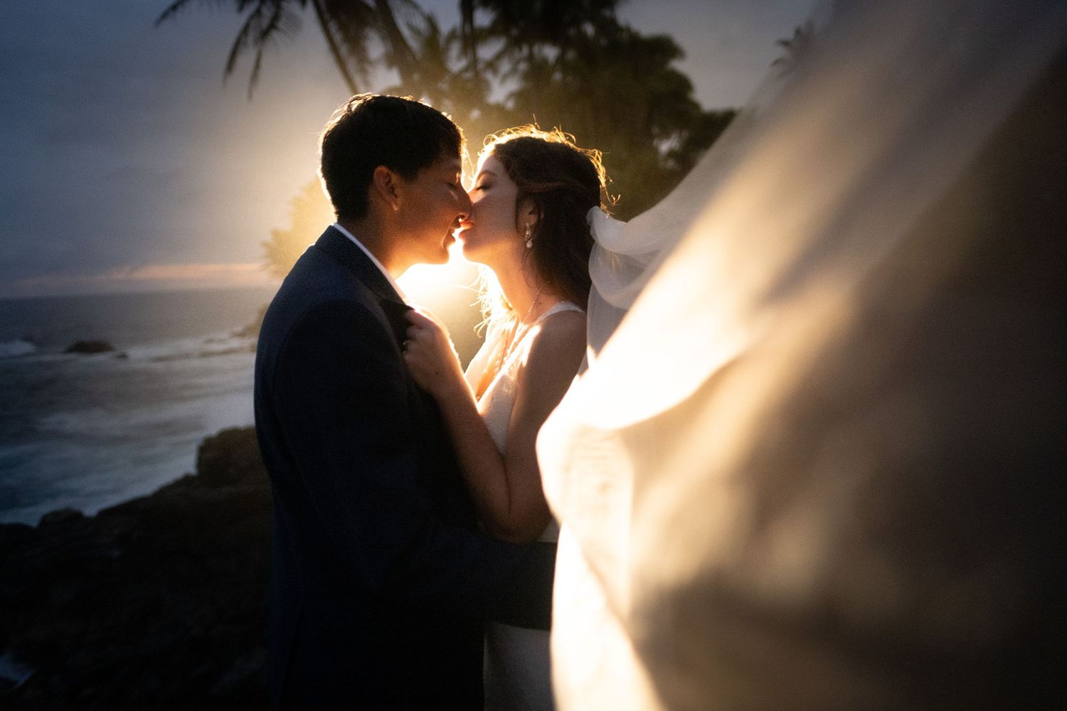 Silhouetted couple embraces during golden hour sunset on a rocky beach at Mauna Lani, Big Island of Hawaii.