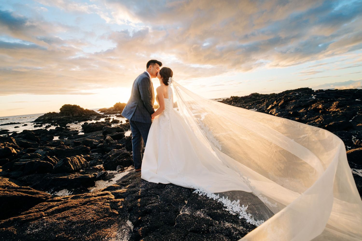 Dramatic sunset wedding portrait on volcanic rocks with bride's veil flowing in the wind at Kikaua Beach, Big Island of Hawaii.