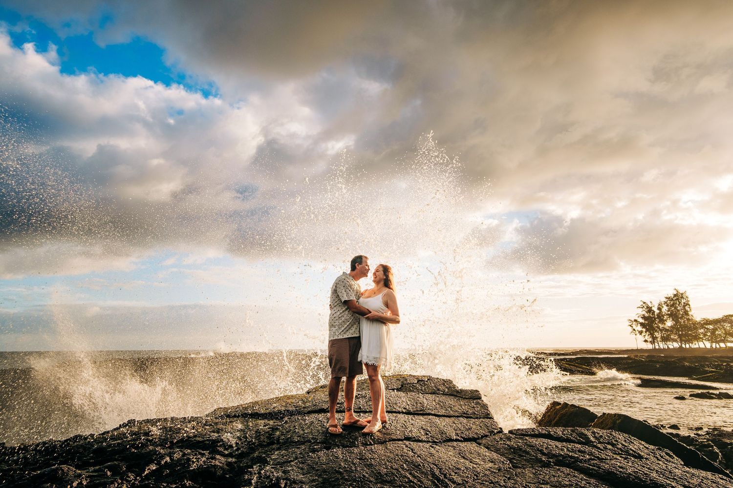 Ocean waves crash dramatically against rocks as people stand on coastal cliffs at sunset at Hilo, Big Island of Hawaii.