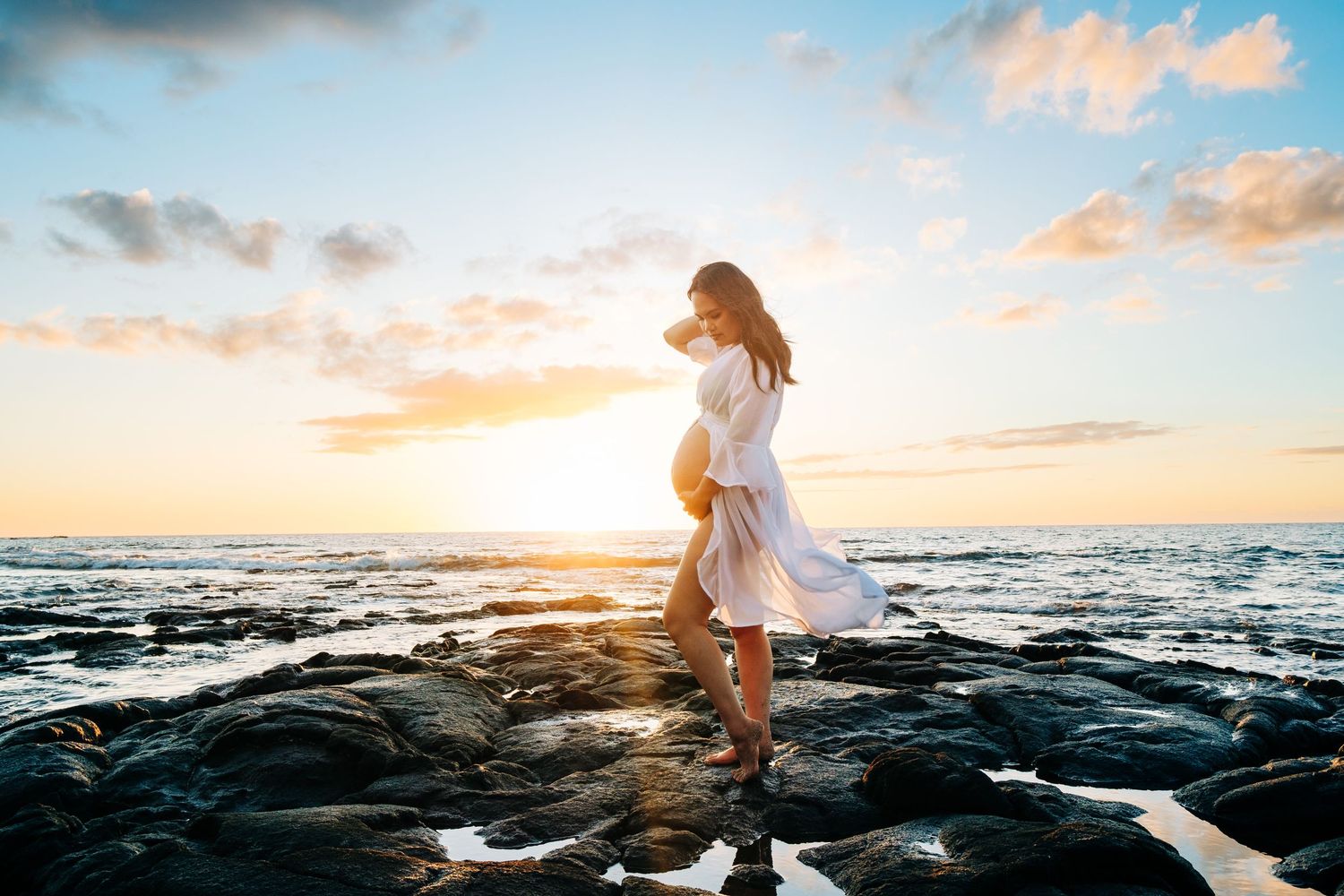 Person in white dress walking on dramatic black lava rocks at sunset by ocean at Kukio Beach, Big Island Hawaii.