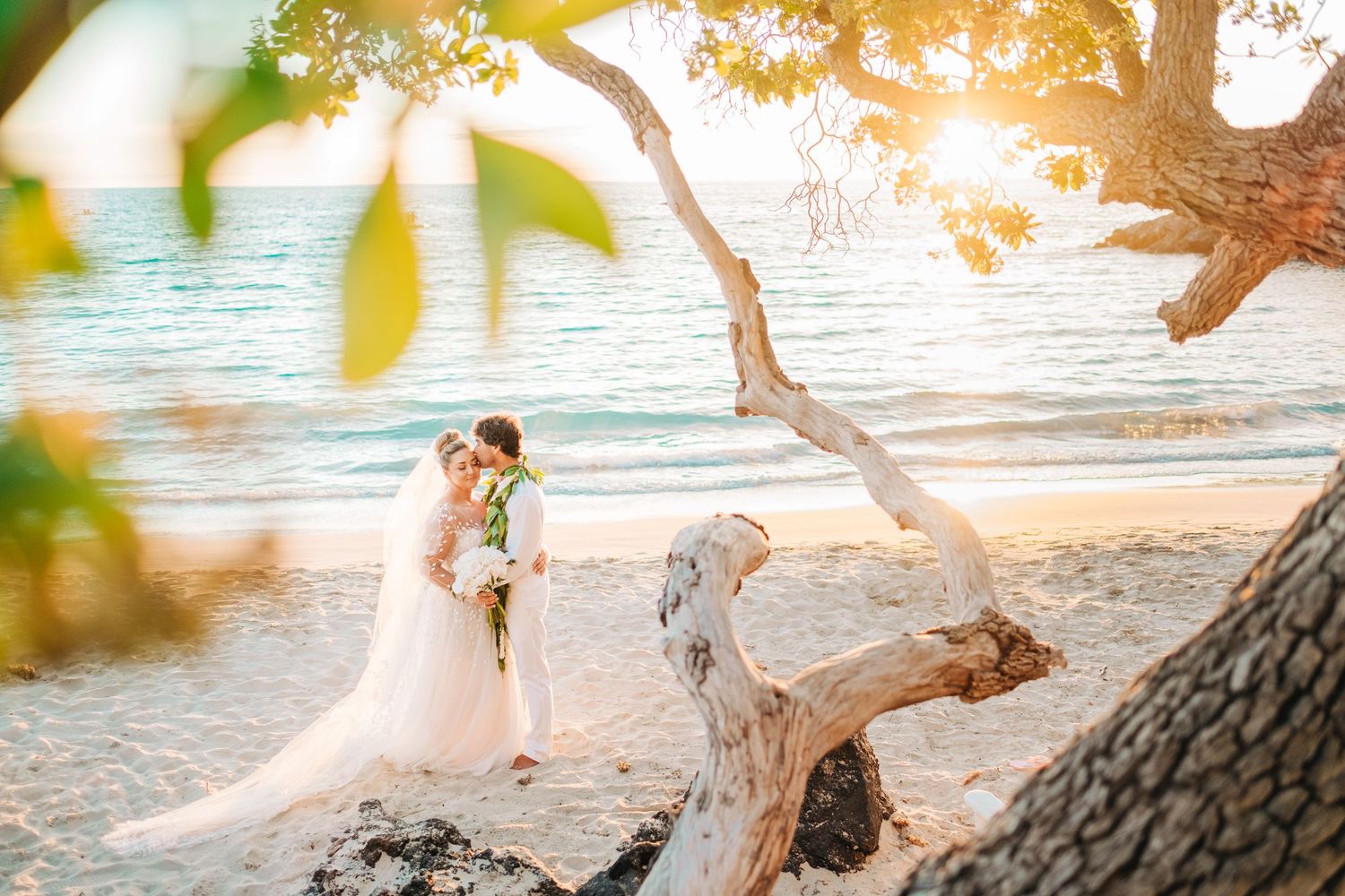 Newlyweds share an intimate moment under a driftwood tree on a tropical beach at sunset. Mauna Kea Beach wedding, Big Island of Hawaii.