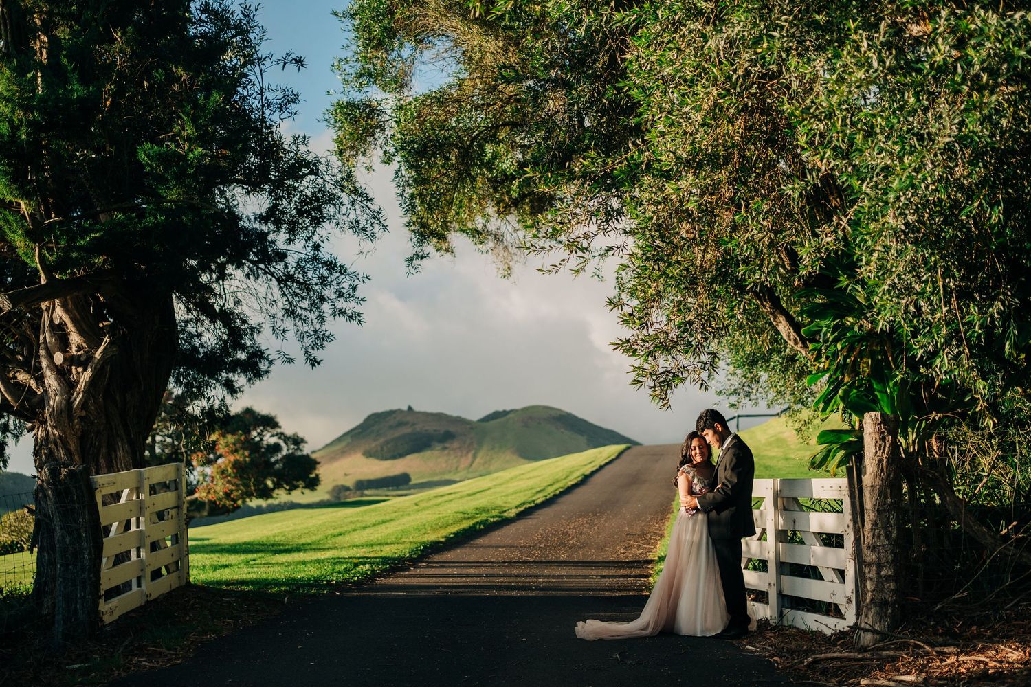 Romantic wedding couple poses along a scenic country road with rolling hills and white fence at sunset at Kahua Ranch, Big Island Hawaii.