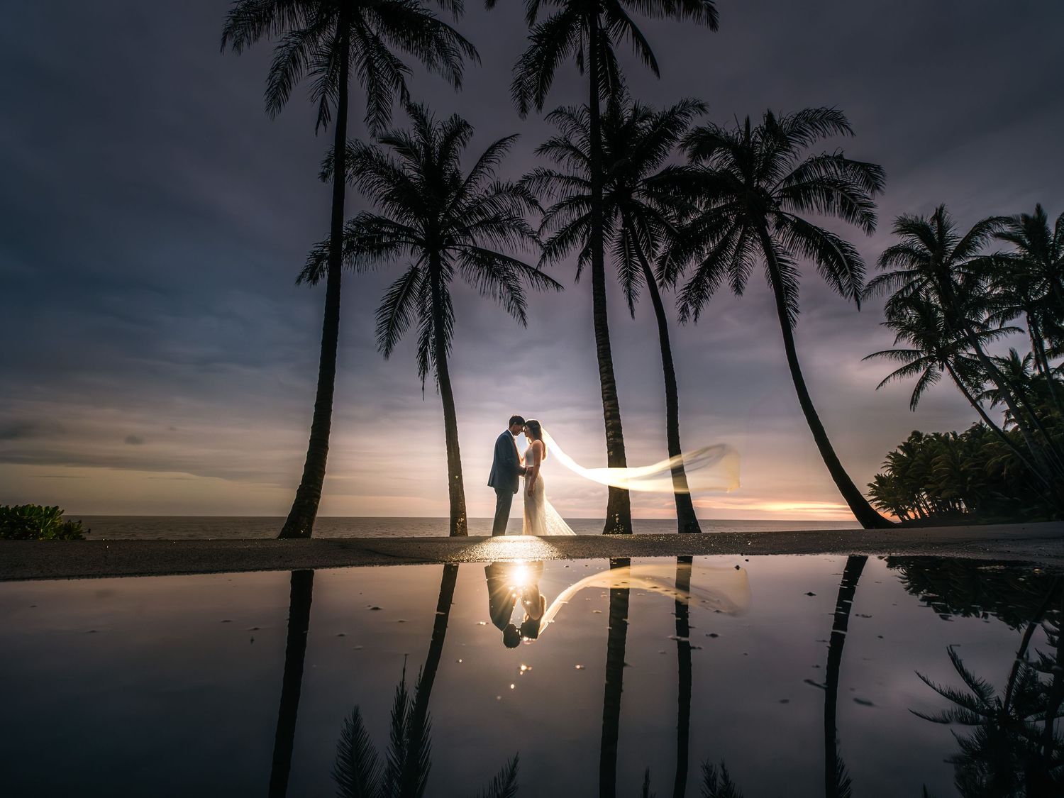 Silhouetted palm trees frame a romantic sunset beach wedding portrait in Hawaii at Mauna Lani, Big Island of Hawaii.