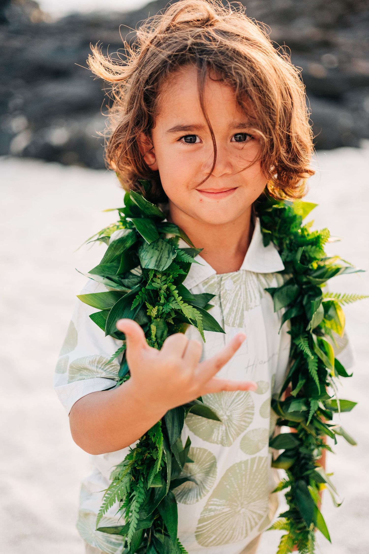 A child wearing a traditional Hawaiian ti leaf lei and white dress poses playfully on a beach at Kona Salt Farm, Big Island of Hawaii.