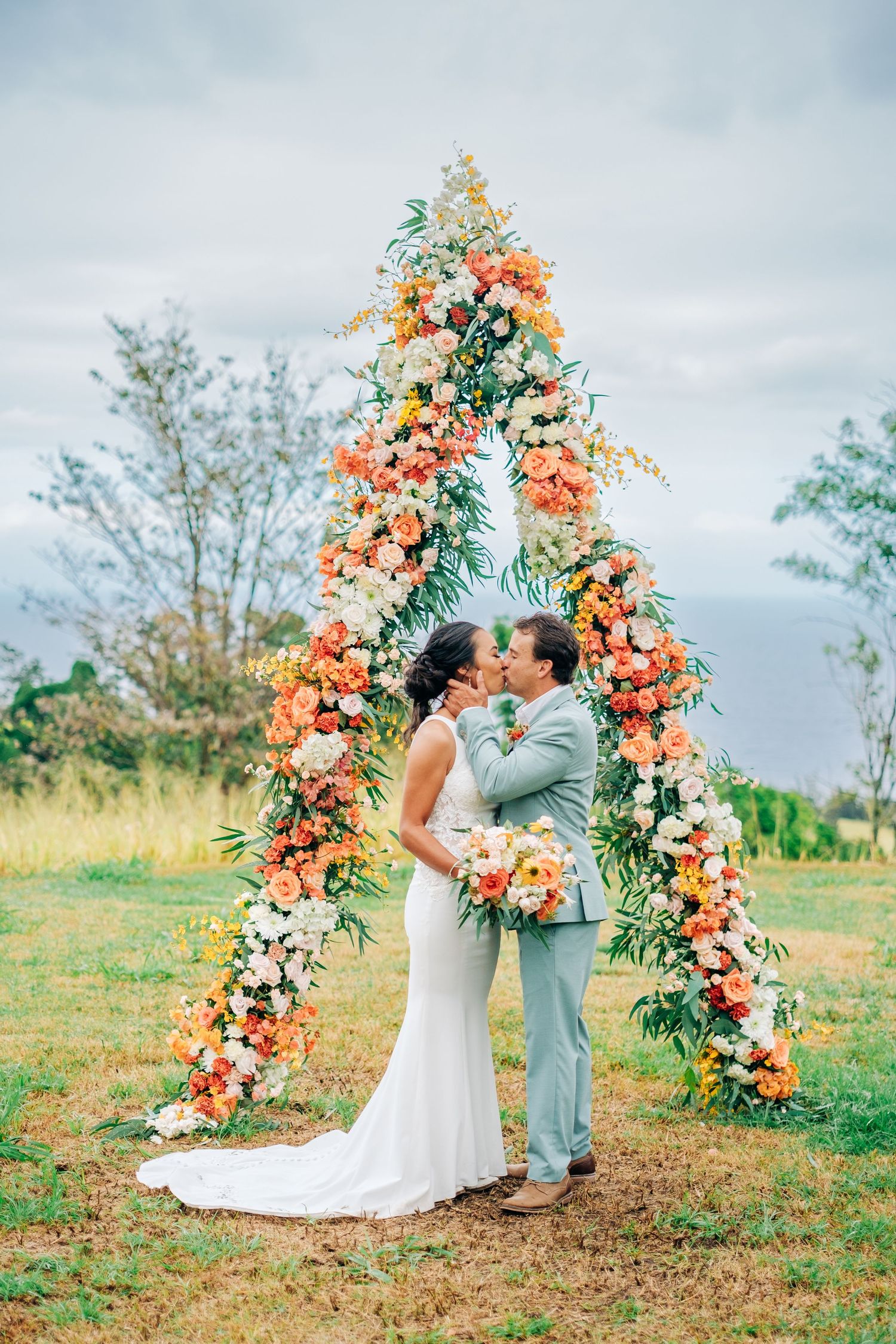 Wedding ceremony under a stunning floral arch with coral and peach blooms in a garden setting on the Big Island of Hawaii.