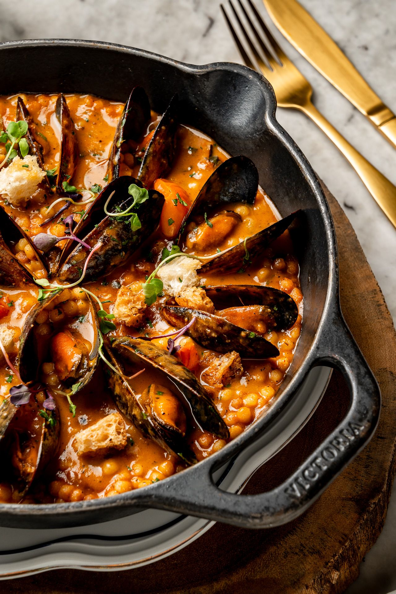 Cast iron pan filled with mussels in rich tomato broth with lentils, garnished with fresh herbs on wooden board.