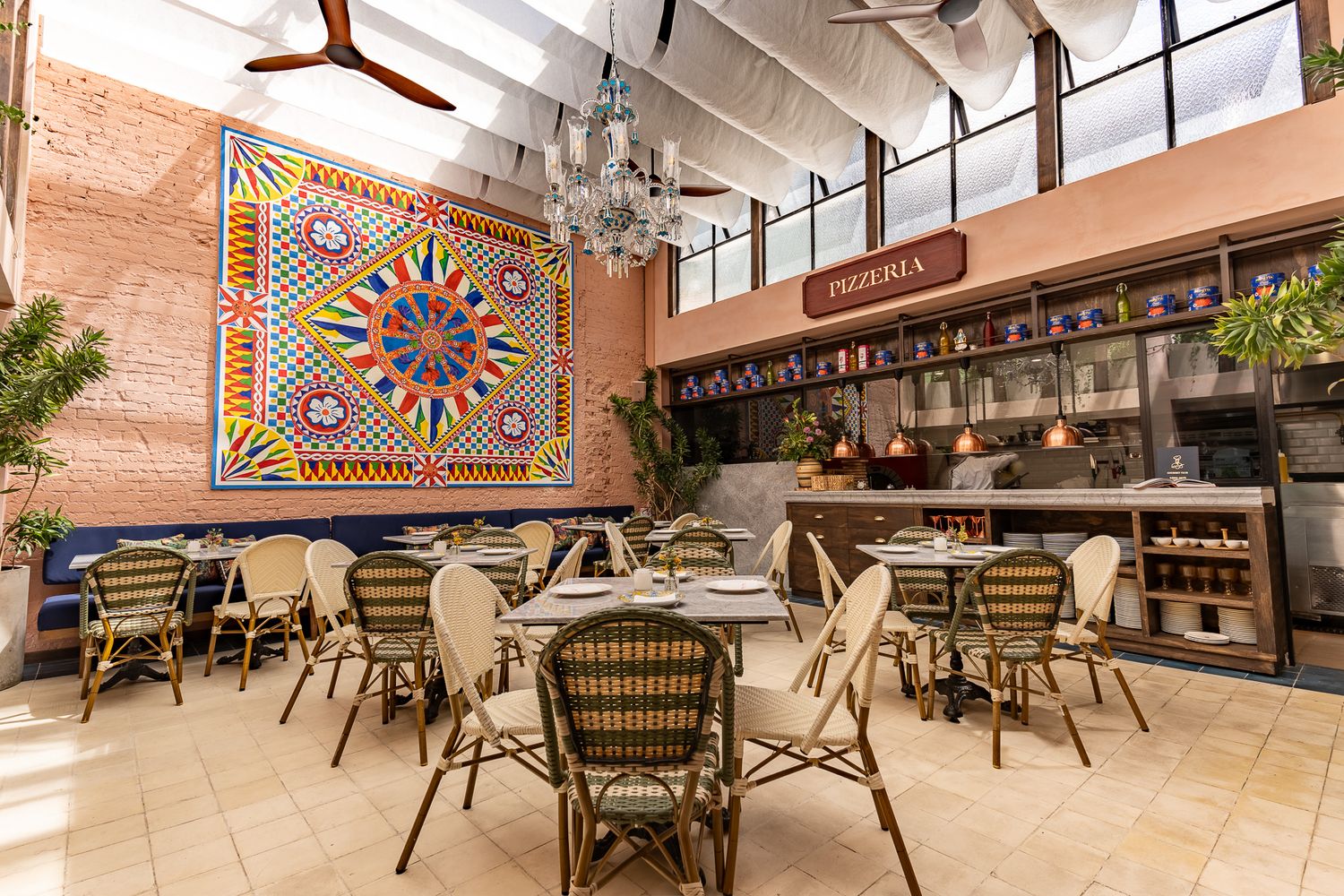 Bright restaurant interior with colorful geometric mural, wicker chairs, chandelier, and open kitchen behind a wooden counter.