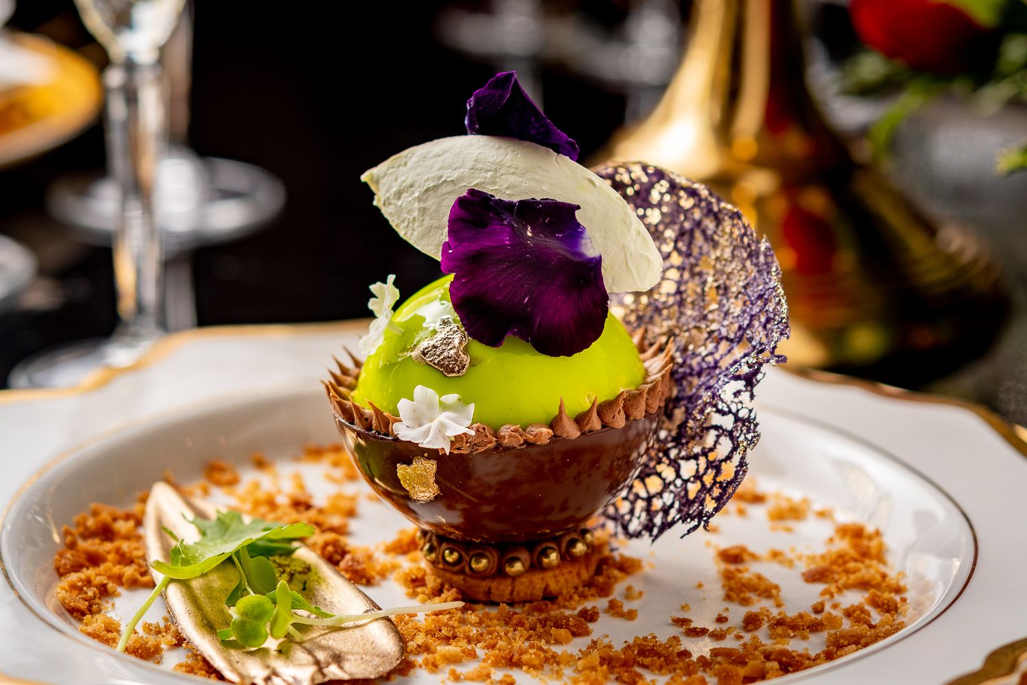 Elegant fine dining dessert with green apple sphere, purple edible flower, chocolate lace, and crumble on white plate.