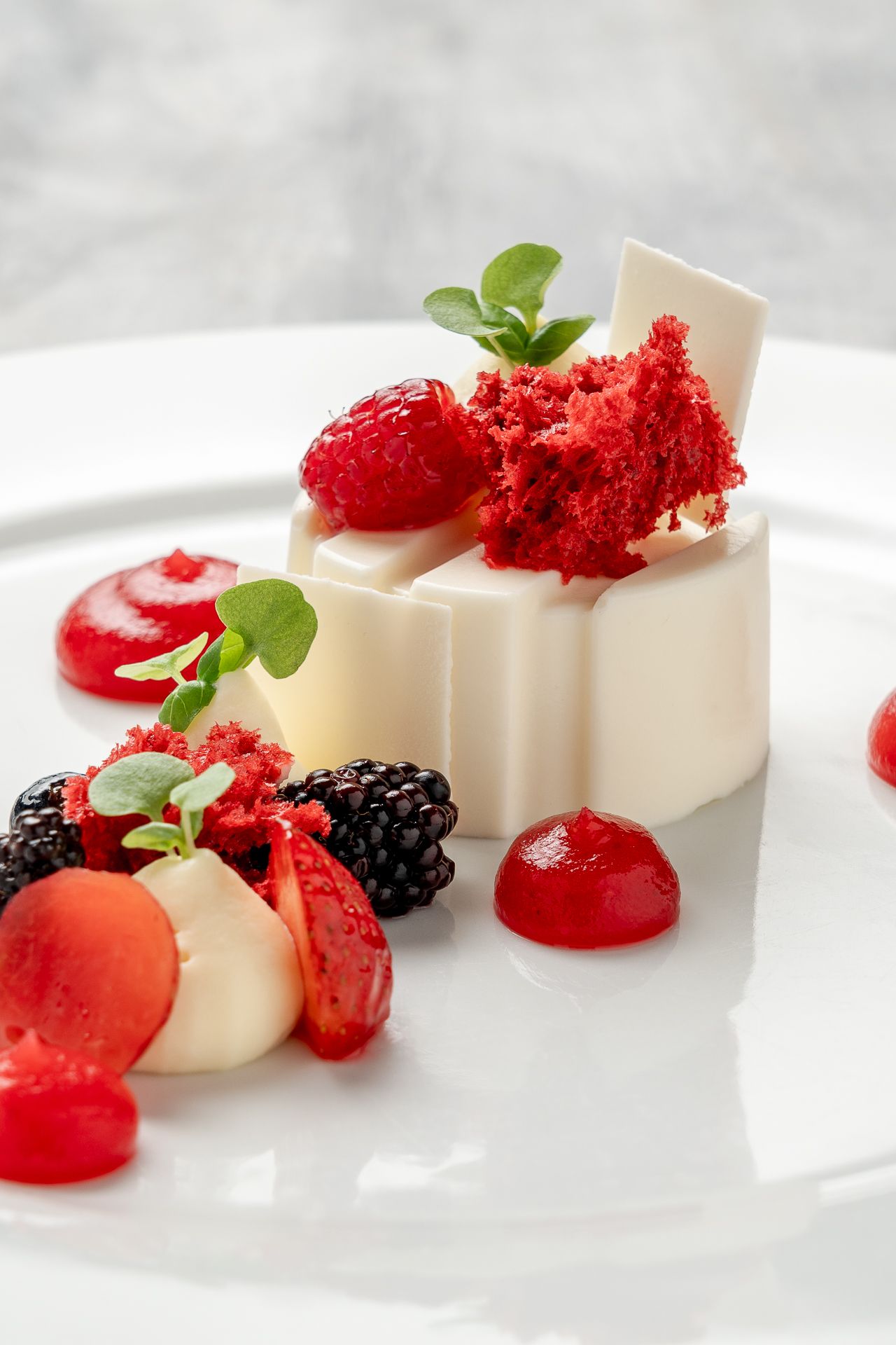 Elegant white chocolate panna cotta dessert with fresh berries, red velvet crumble, and mint garnish on a white plate.
