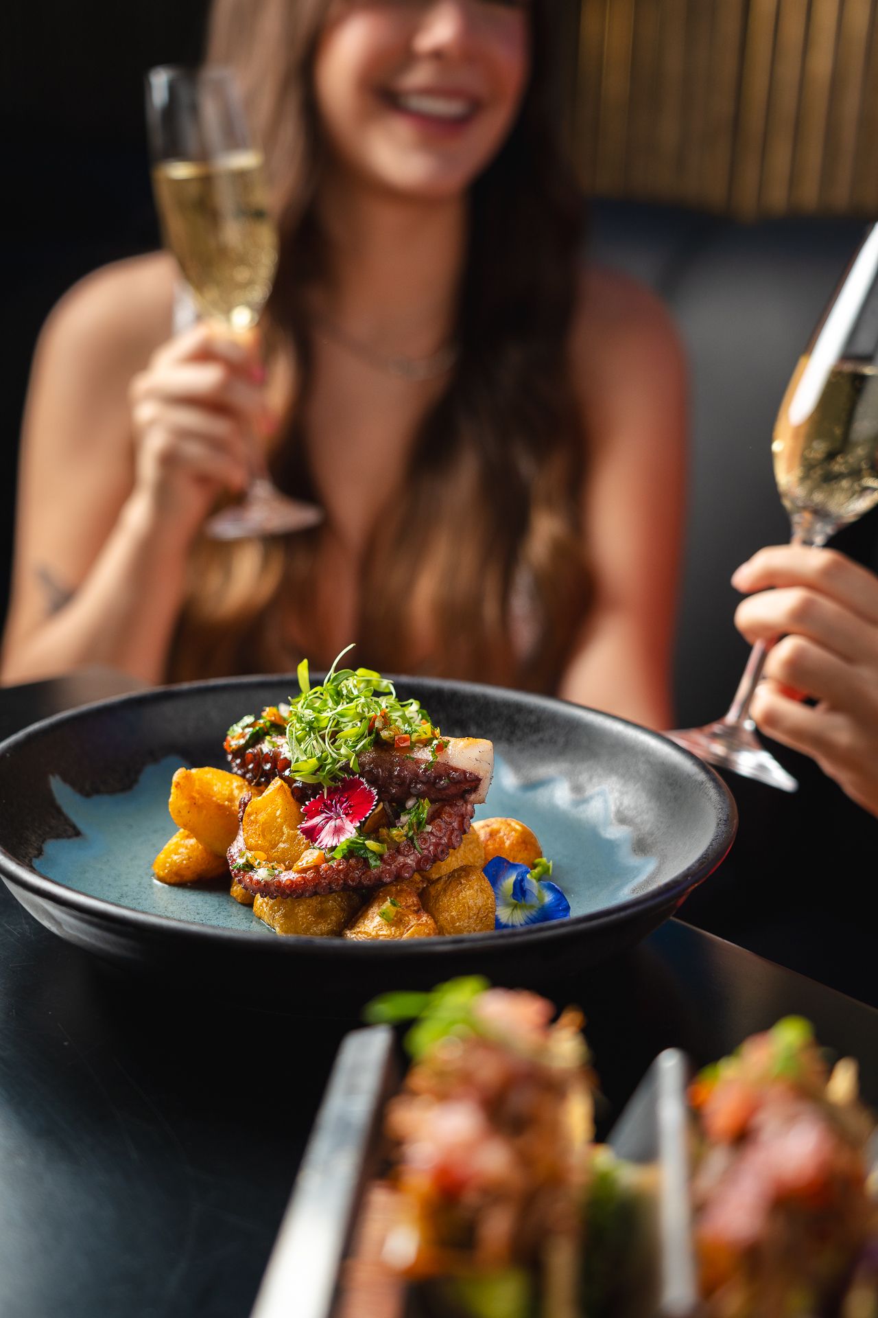 Elegant dining scene with gourmet octopus dish on dark plate, woman holding champagne glass in background.