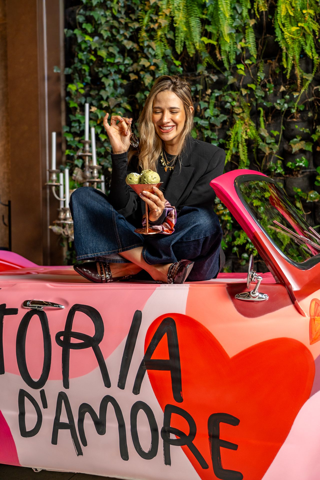 Woman sitting on a pink car painted with 'Storia D'Amore', holding an ice cream cocktail, smiling outdoors.