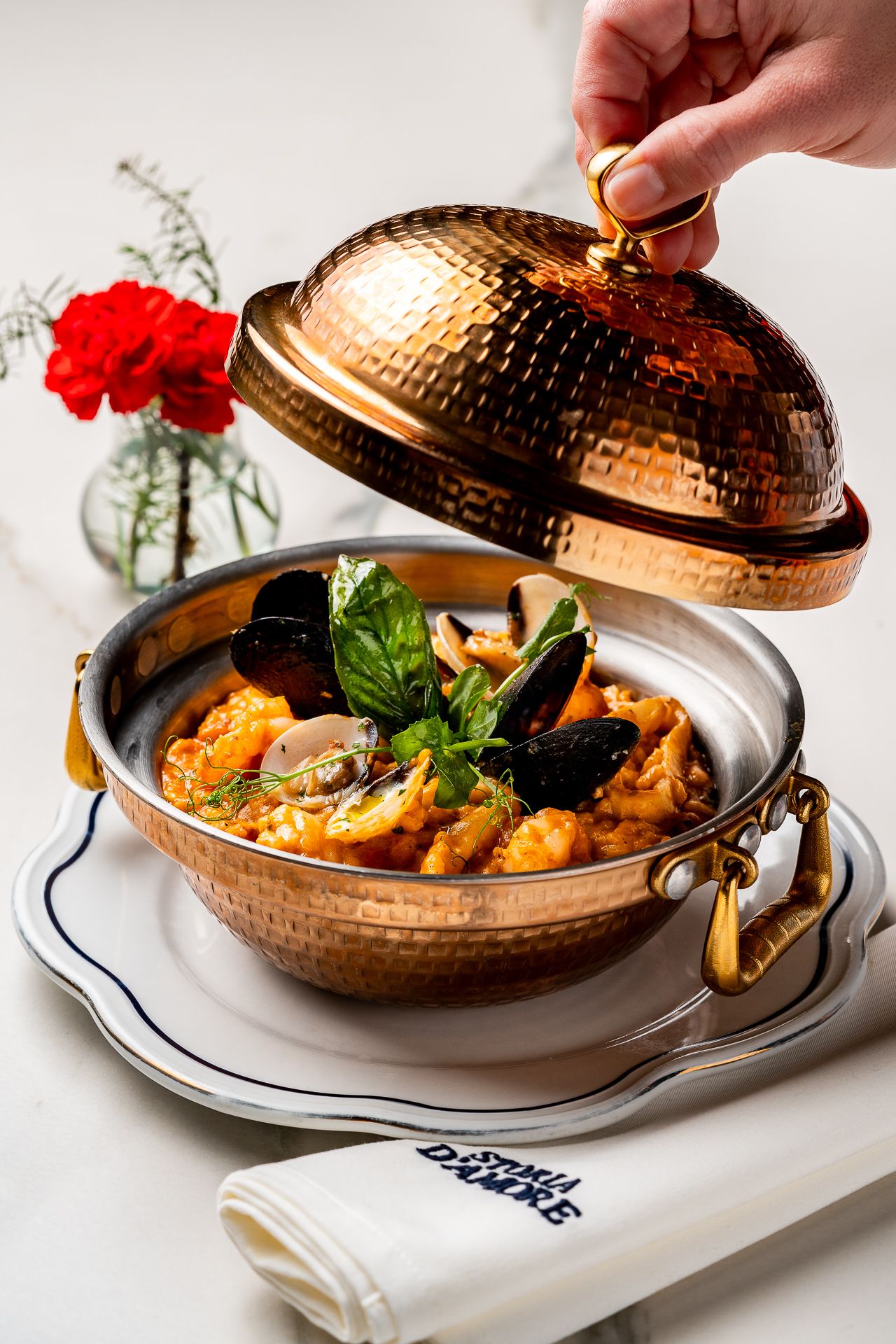 Copper pot with lid being lifted to reveal seafood stew with mussels and shrimp, garnished with fresh basil.