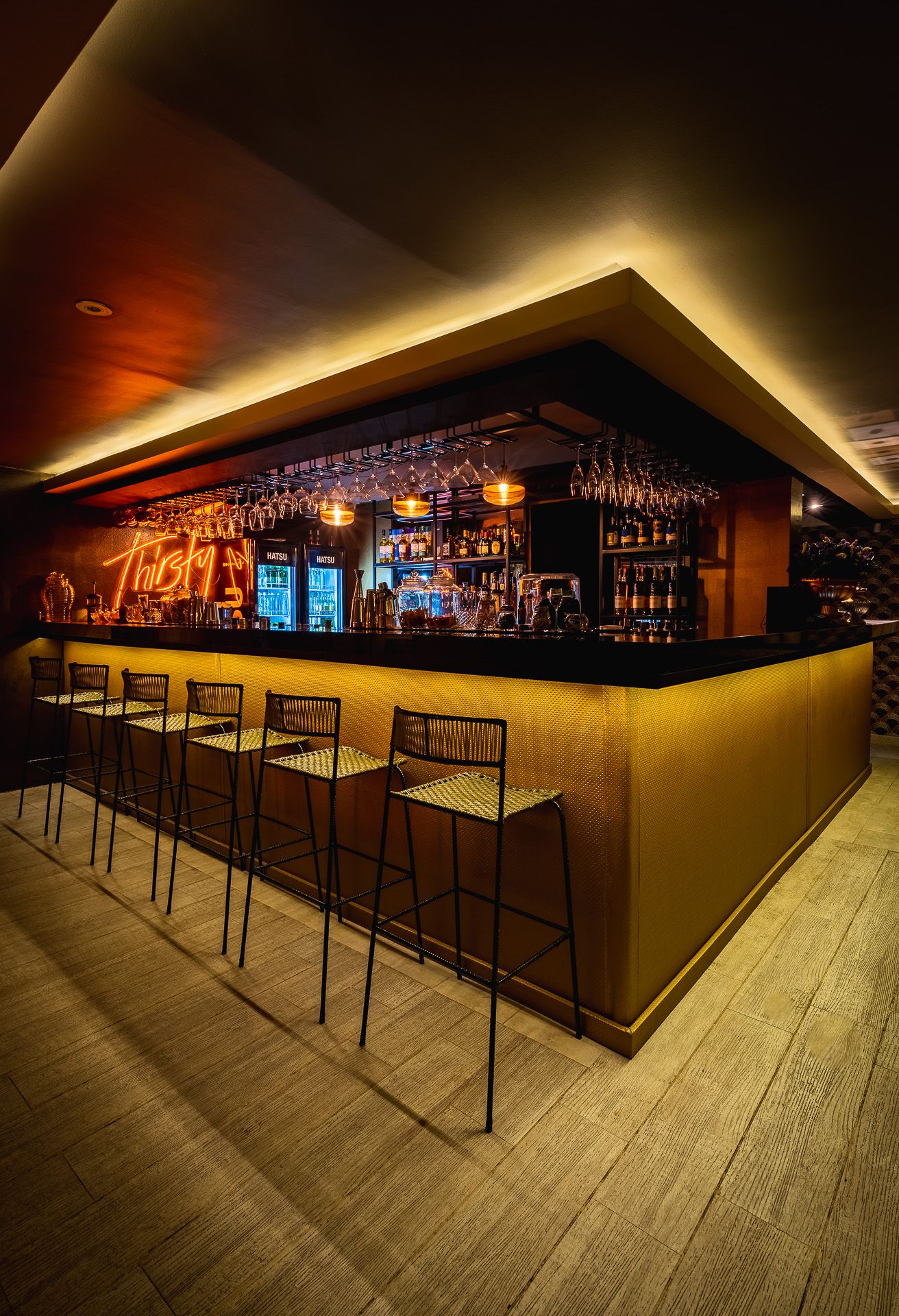 Stylish bar interior with yellow-lit counter, bar stools, hanging wine glasses, and warm ambient lighting.