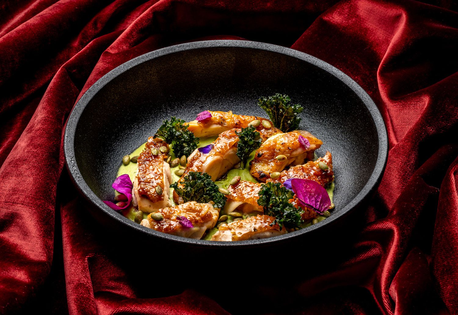 Elegant chicken dish with broccoli and purple flowers in a dark bowl on red silk fabric.