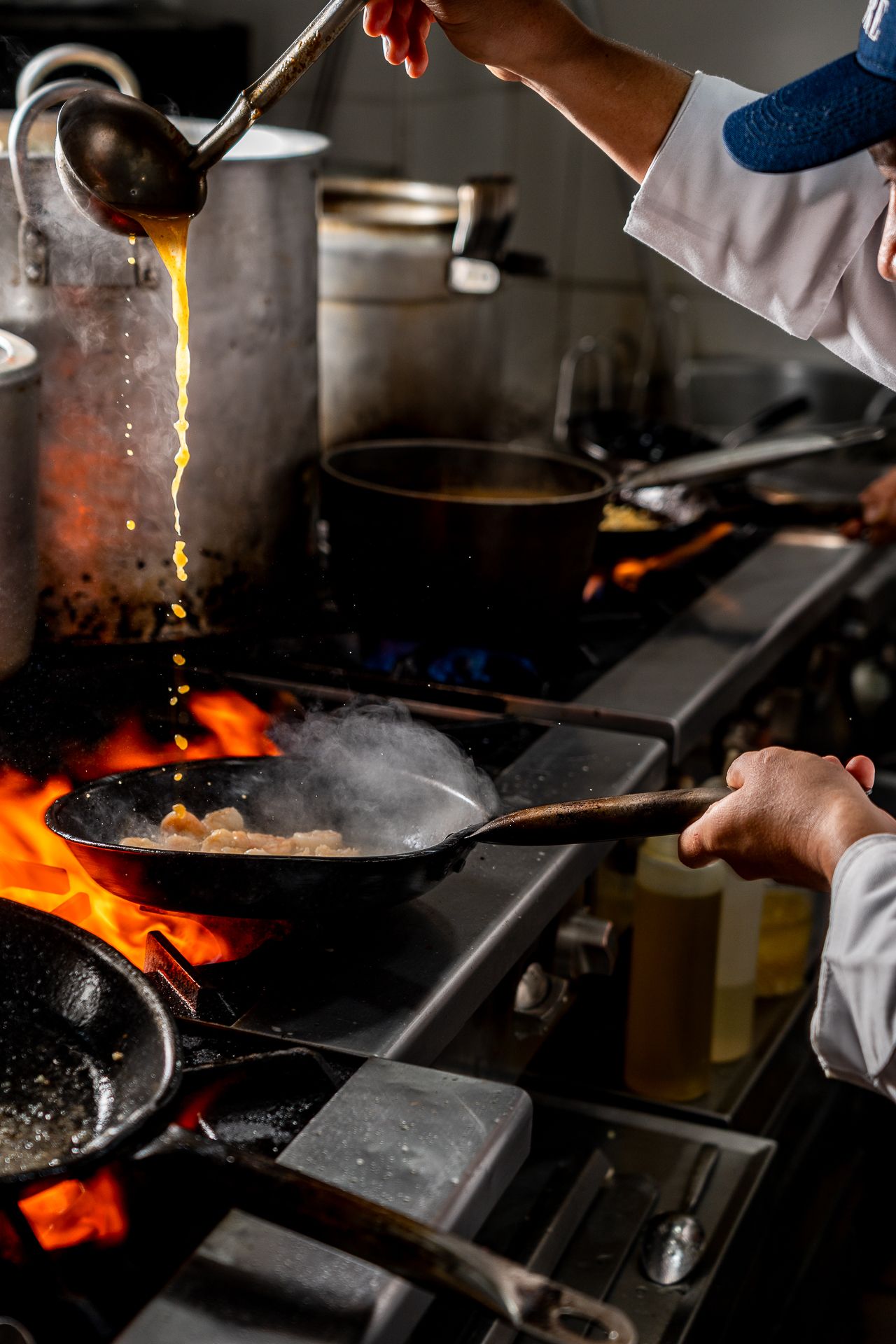 Chef cooking with open flames on a commercial stove, pouring sauce into a sizzling pan in a professional kitchen.