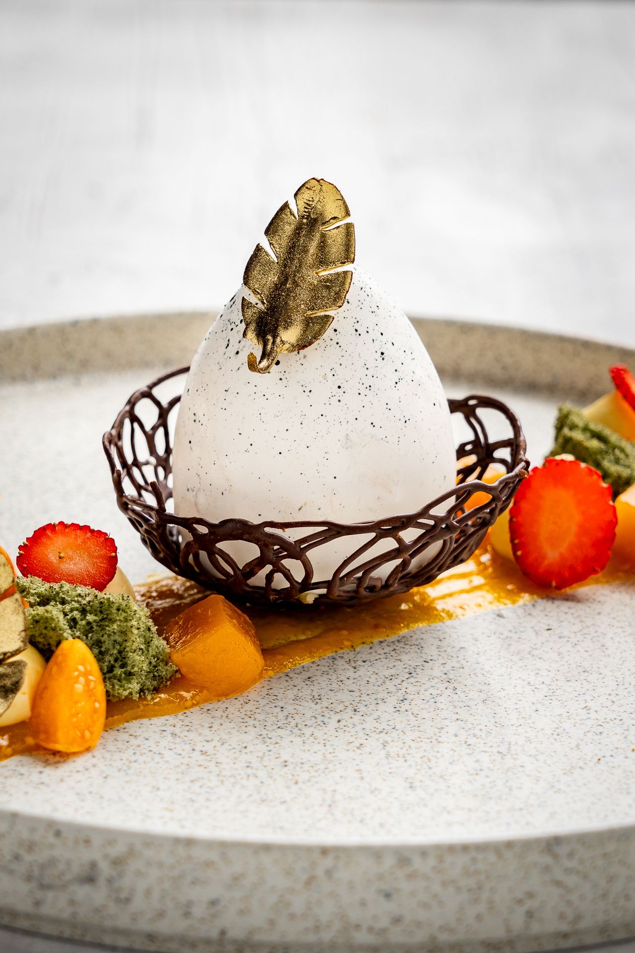 Elegant dessert with white mousse dome in chocolate lace basket, topped with gold leaf, served with fresh strawberries and mango.