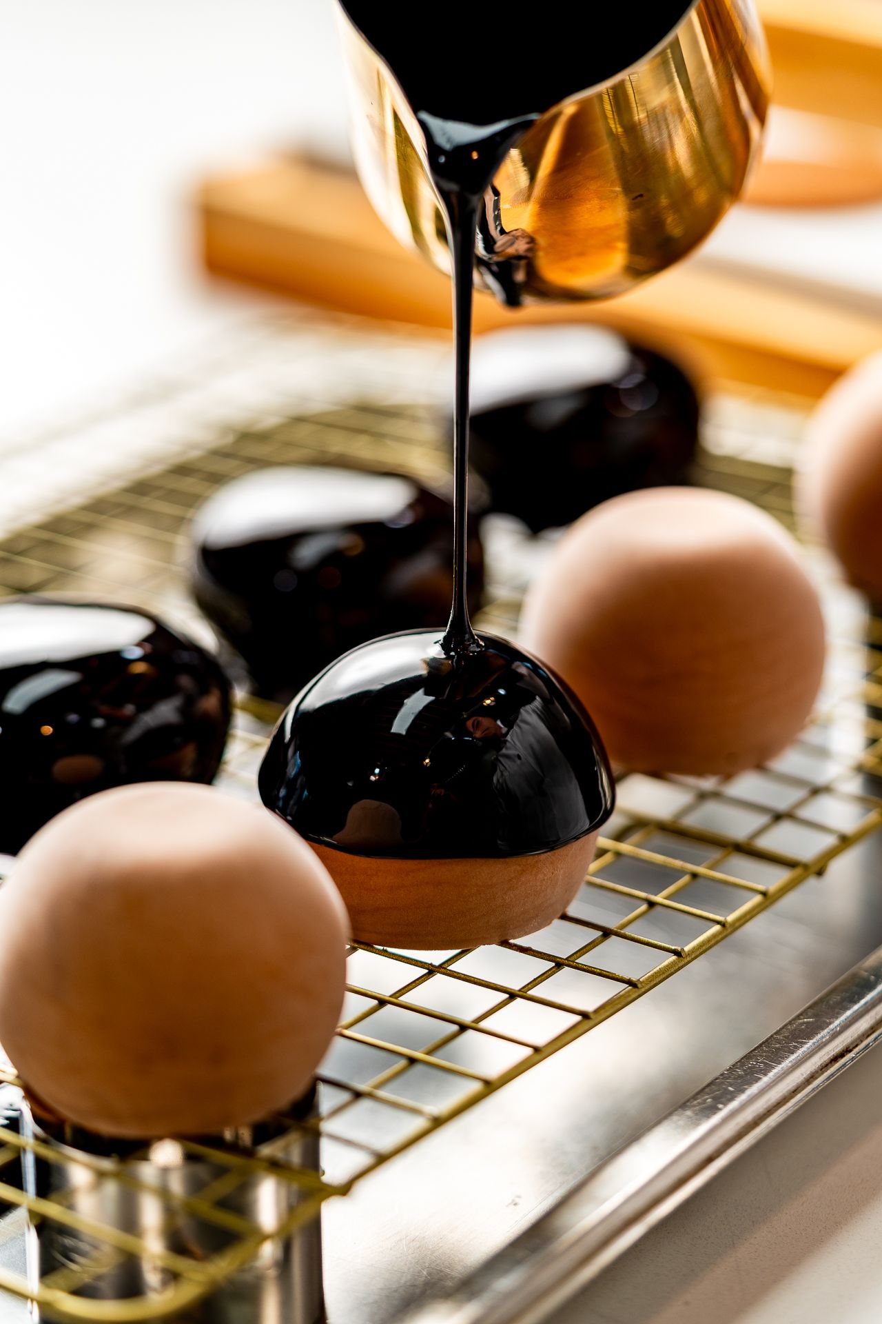 Dark chocolate glaze being poured over round chocolate bonbons on a gold wire cooling rack.