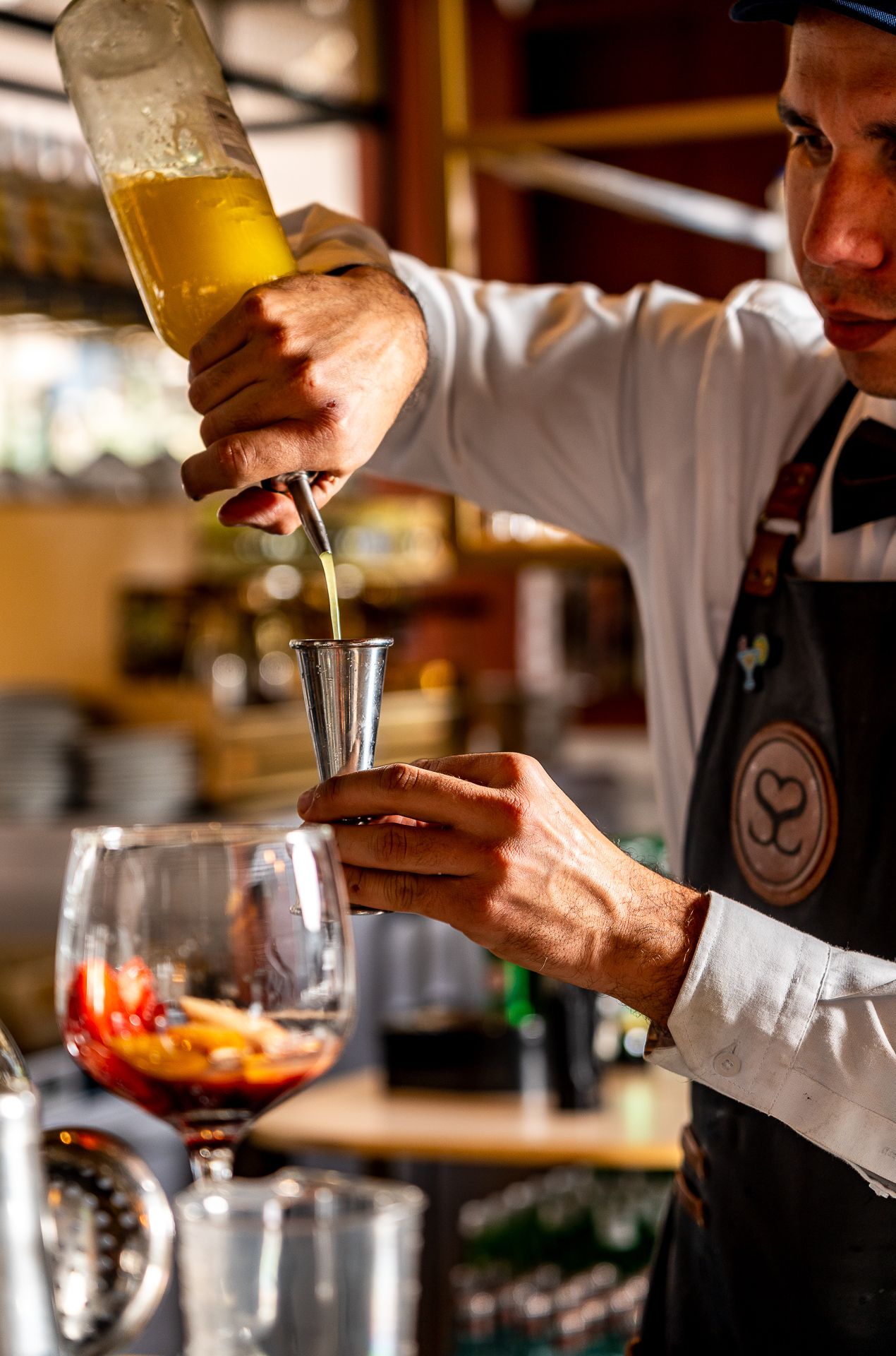 Bartender in black apron pouring yellow liquid into a jigger while preparing a cocktail at a bar.