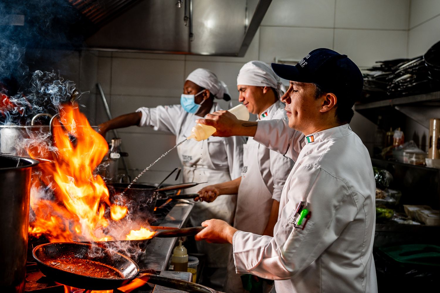 Chefs in white uniforms cooking with open flames in a busy restaurant kitchen.