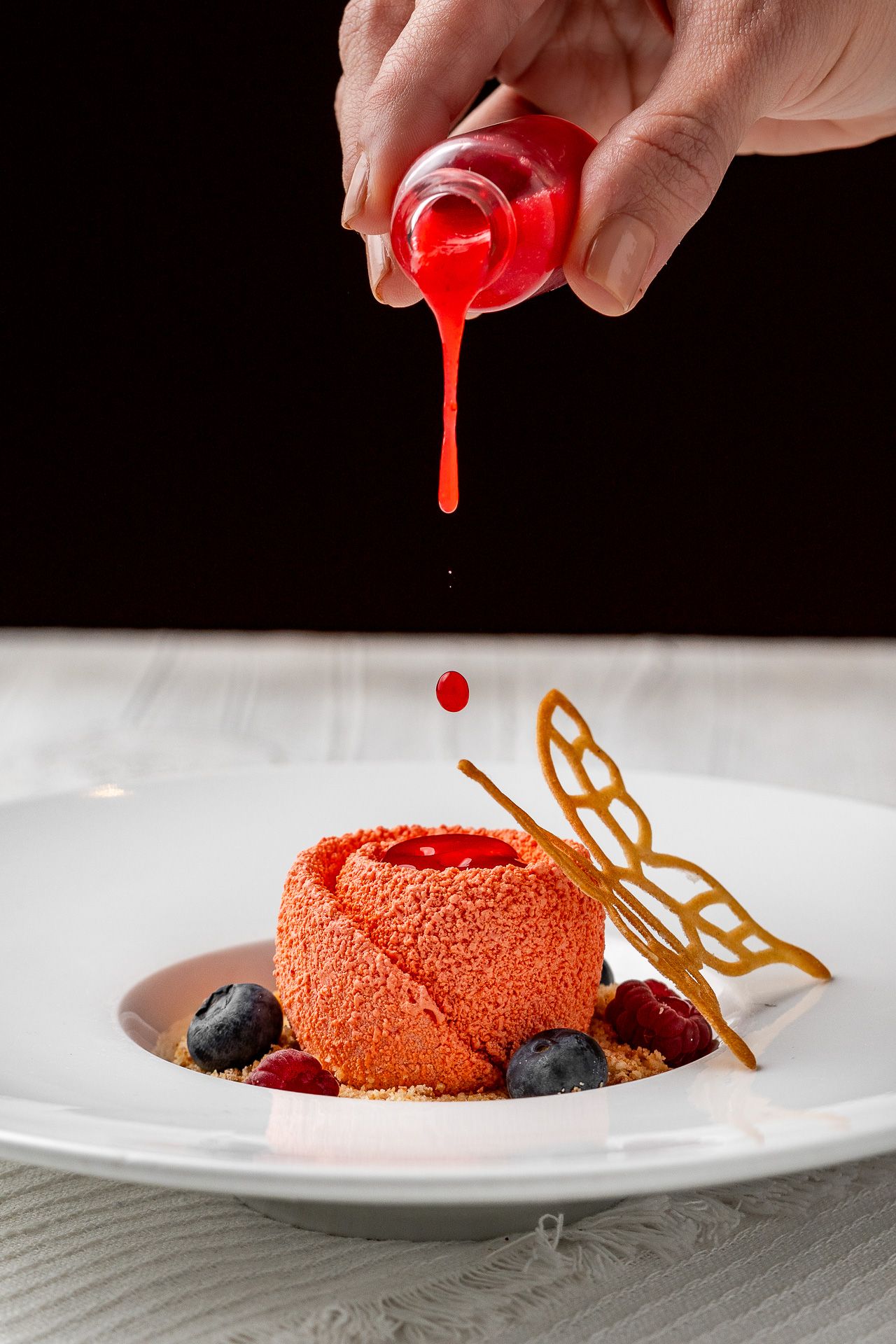 Hand pouring red berry sauce over elegant pink mousse dessert with blueberries and golden butterfly decoration on white plate.