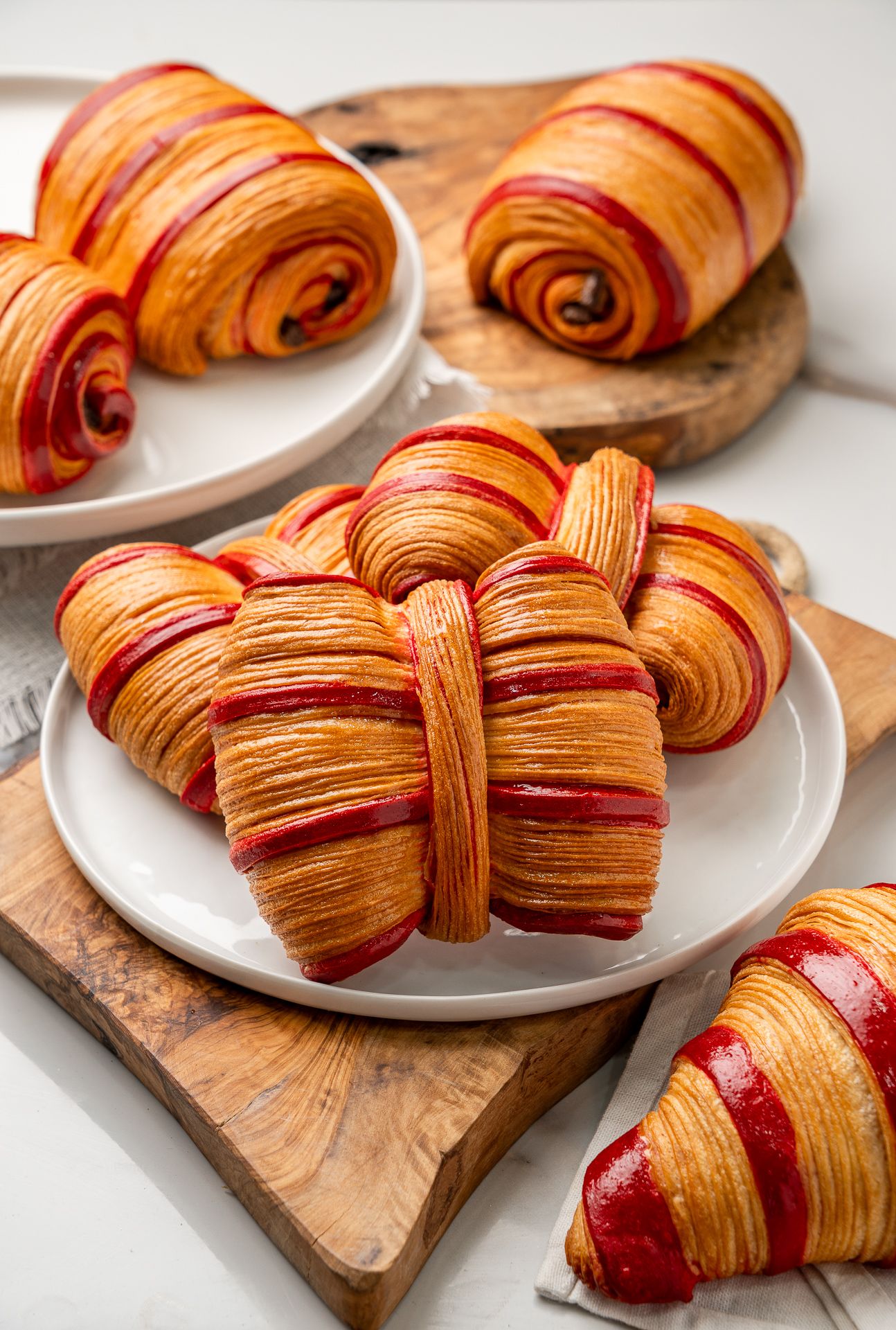 Flaky red-striped croissants on white plates and wooden boards, showcasing vibrant layered pastry with crimson swirls.