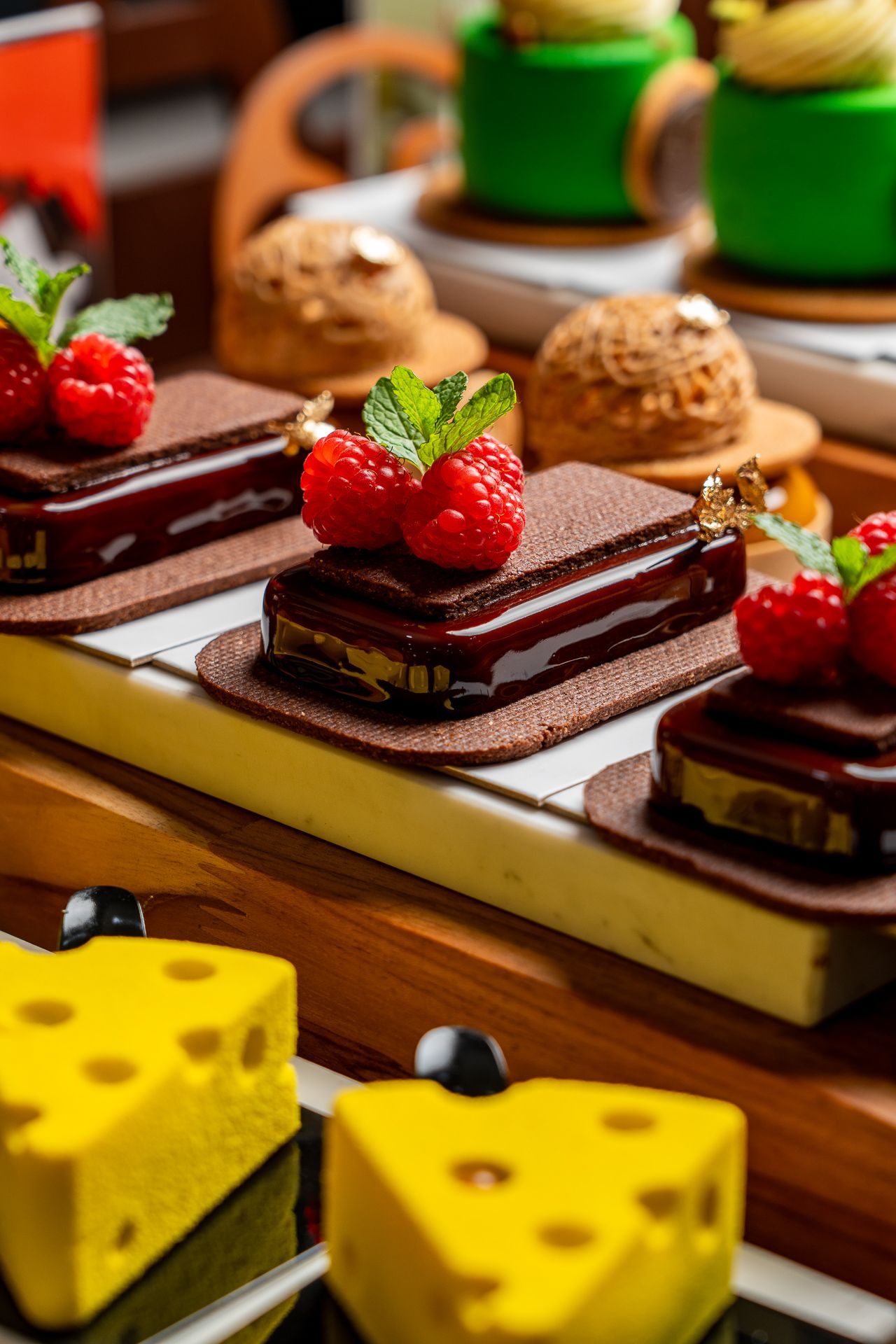 Elegant chocolate desserts topped with fresh raspberries and mint, displayed alongside colorful pastries on a wooden table.