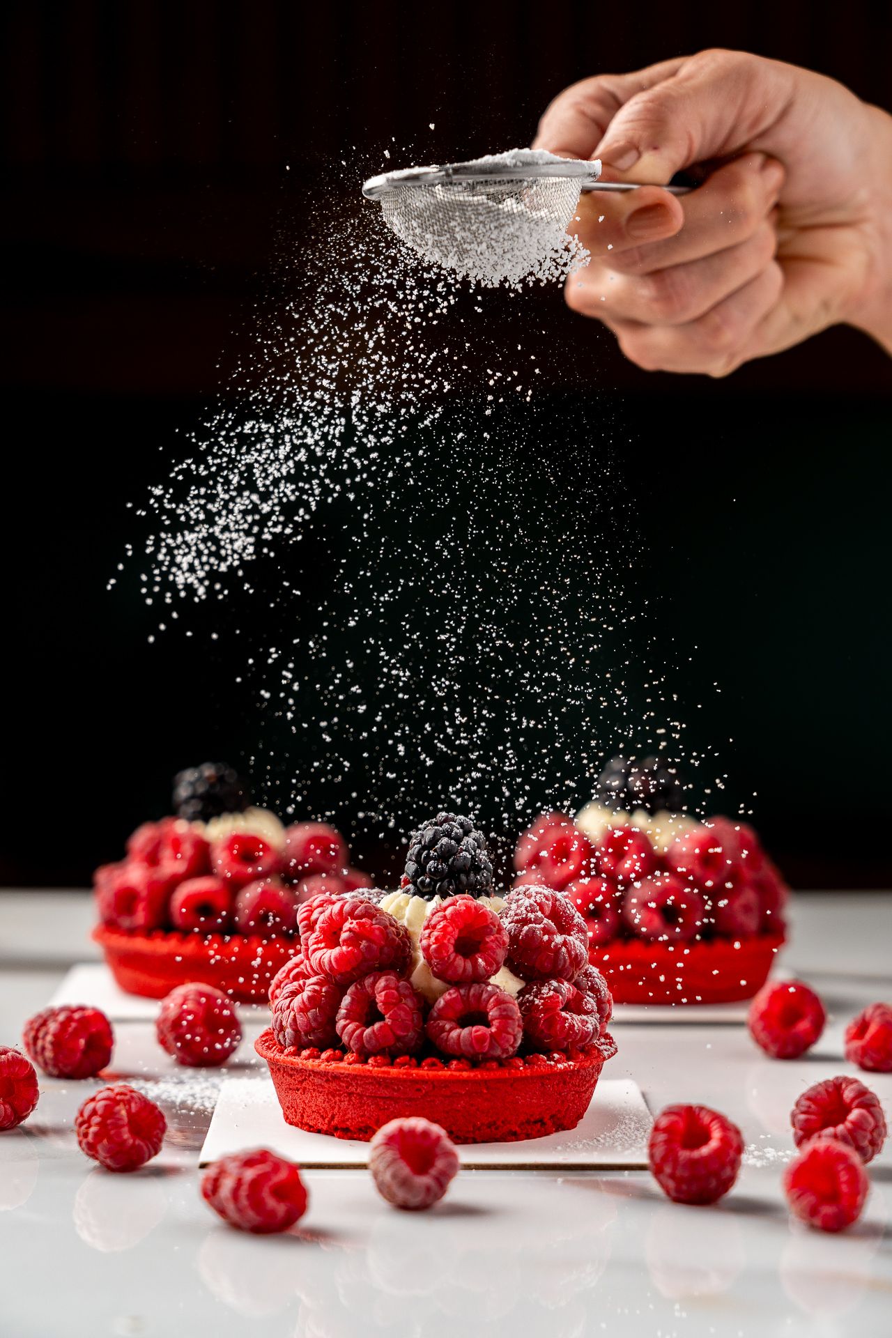 Hand dusting powdered sugar over red raspberry tarts on white surface with fresh raspberries scattered around.